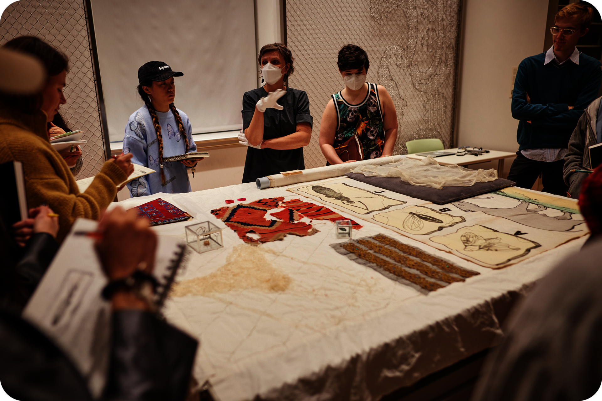 Several people standing around a table displaying various textiles. A person in the center wearing gloves gestures towards another person, while others stare at the textiles on the table. 