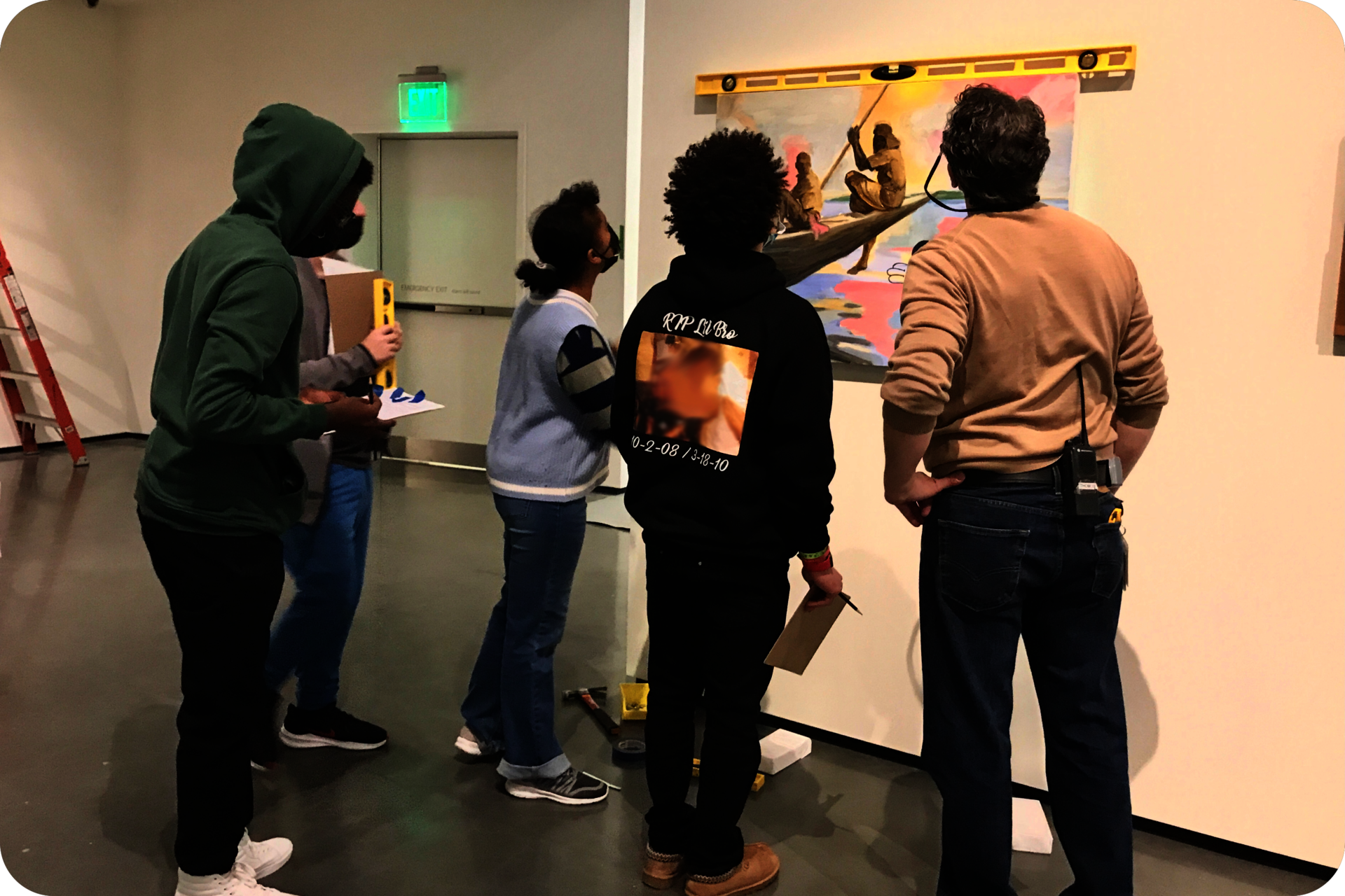 5 people standing around and looking at a painting hung on a wall. A level sits on top of the painting, and several people are holding various tools. 