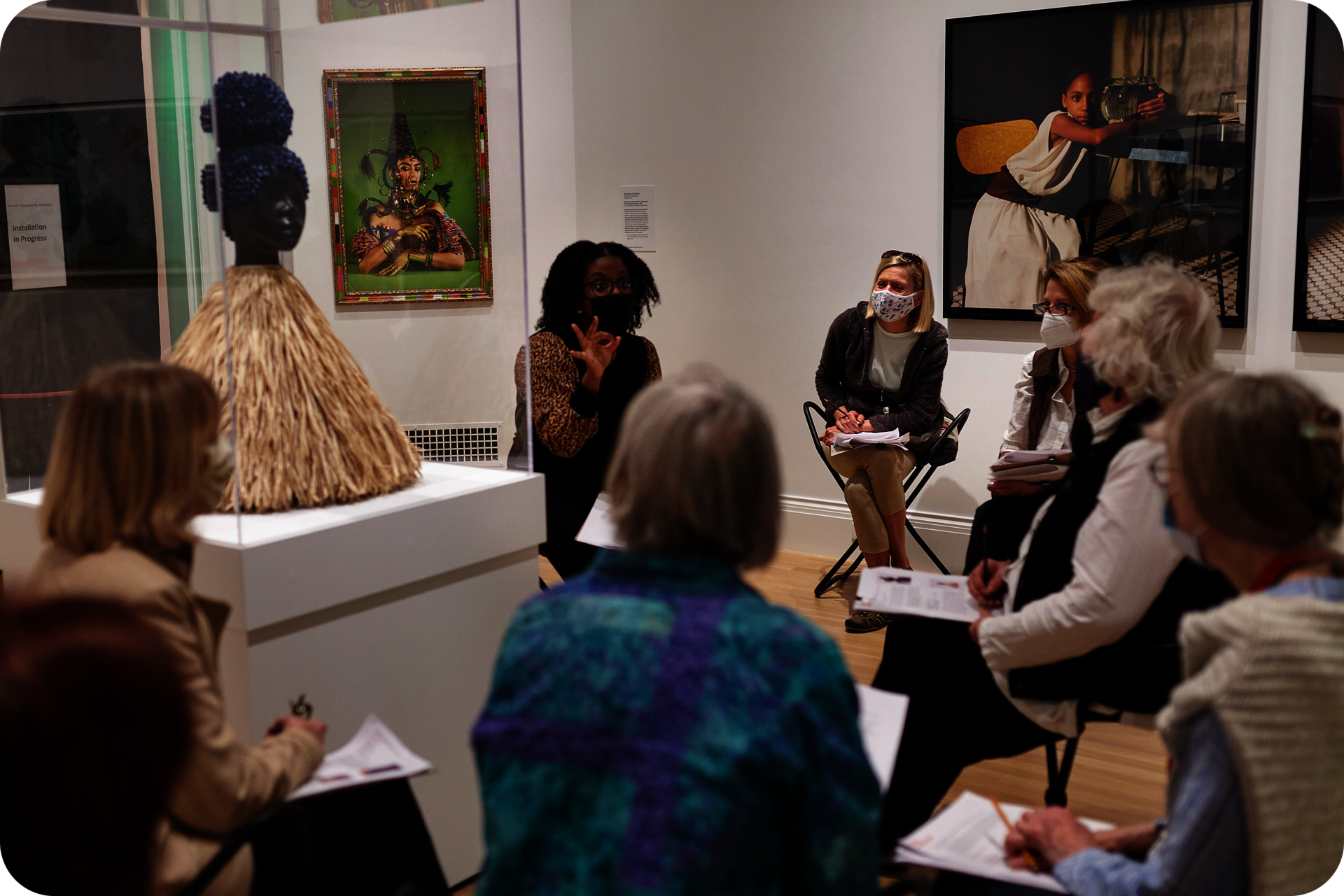 Group of people holding papers sitting around a bust sculpture encased in glass. A person in the center gestures the number three. The wall behind them features framed photographs.