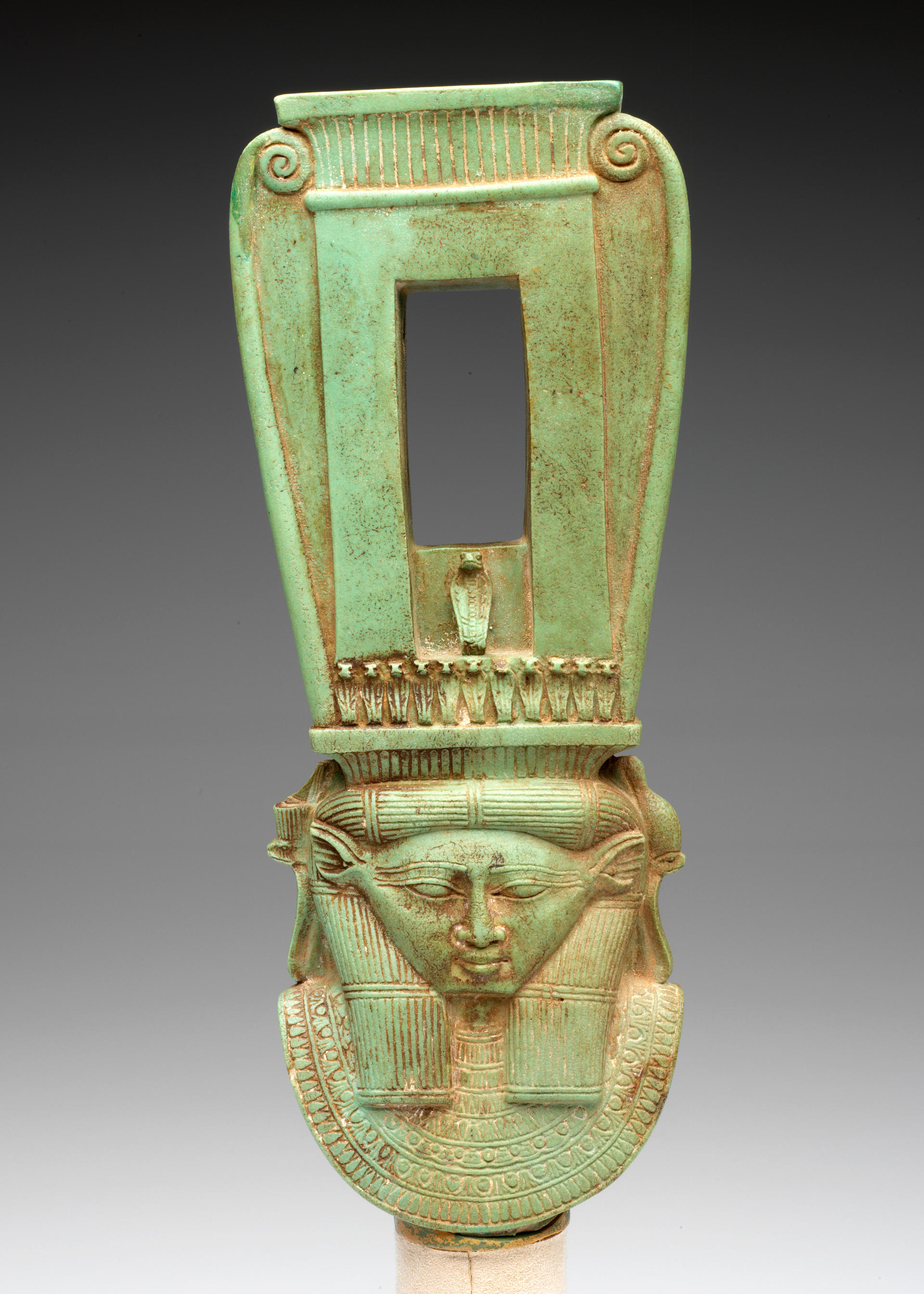 Front view of a long jade-green sculpture of a collared figure wearing a tall headdress, which has a rectangular opening and swirls on the outer edges forming a curved top.