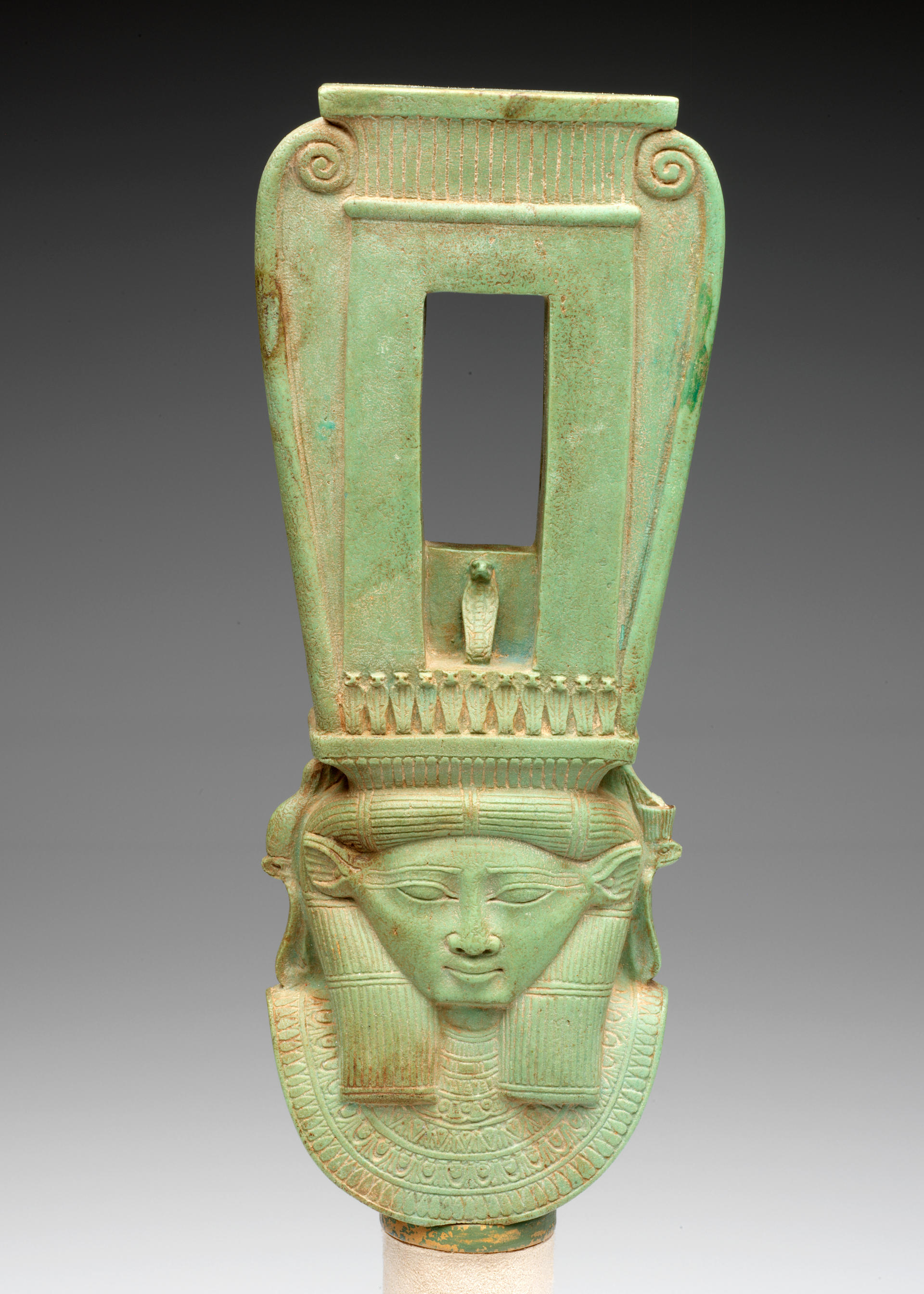 Front view of a long jade-green sculpture of a collared figure wearing a tall headdress, which has a rectangular opening and swirls on the outer edges forming a curved top.