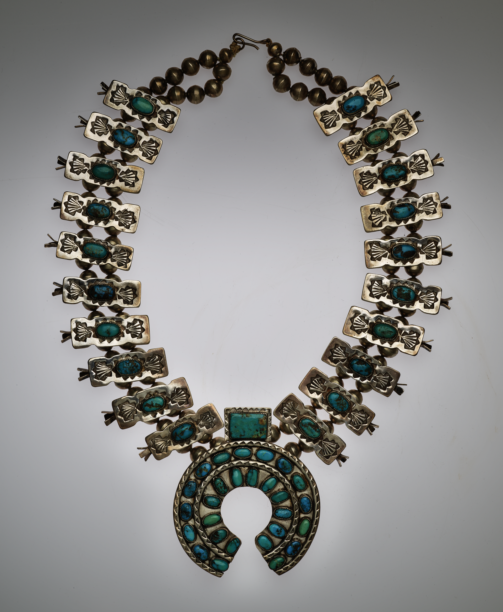 Silver and turquoise necklace. The chain features turquoise stones set within silver rectangular ornaments threaded into two rows of beaded chains. The central pendant is a turquoise stone-set horseshoe shape. 