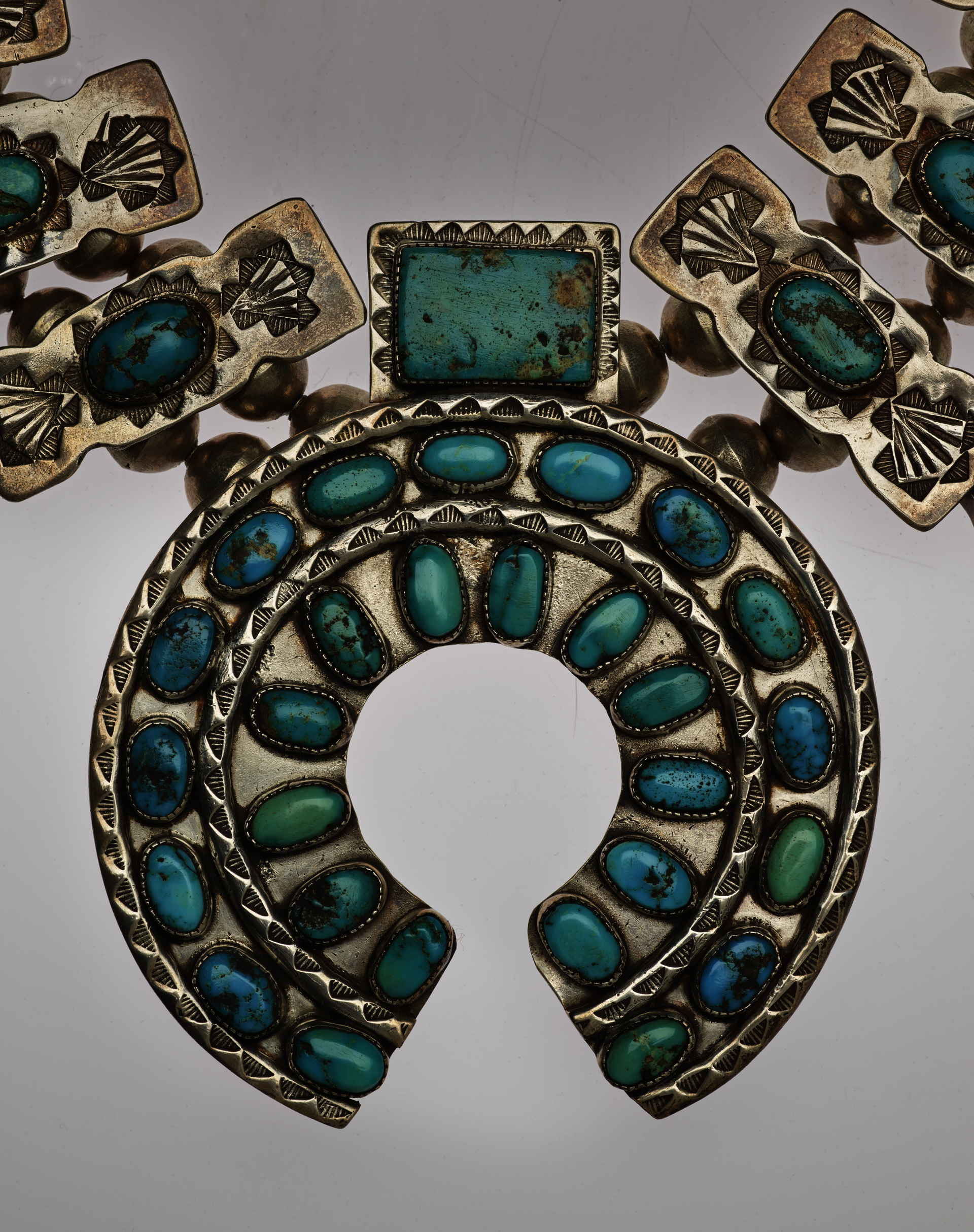 Detail-view of a horseshoe-shaped pendant of a turquoise and silver necklace. The turquoise stones are set in two concentric circles with decorative silver borders with a rectangular stone-setting atop it.
