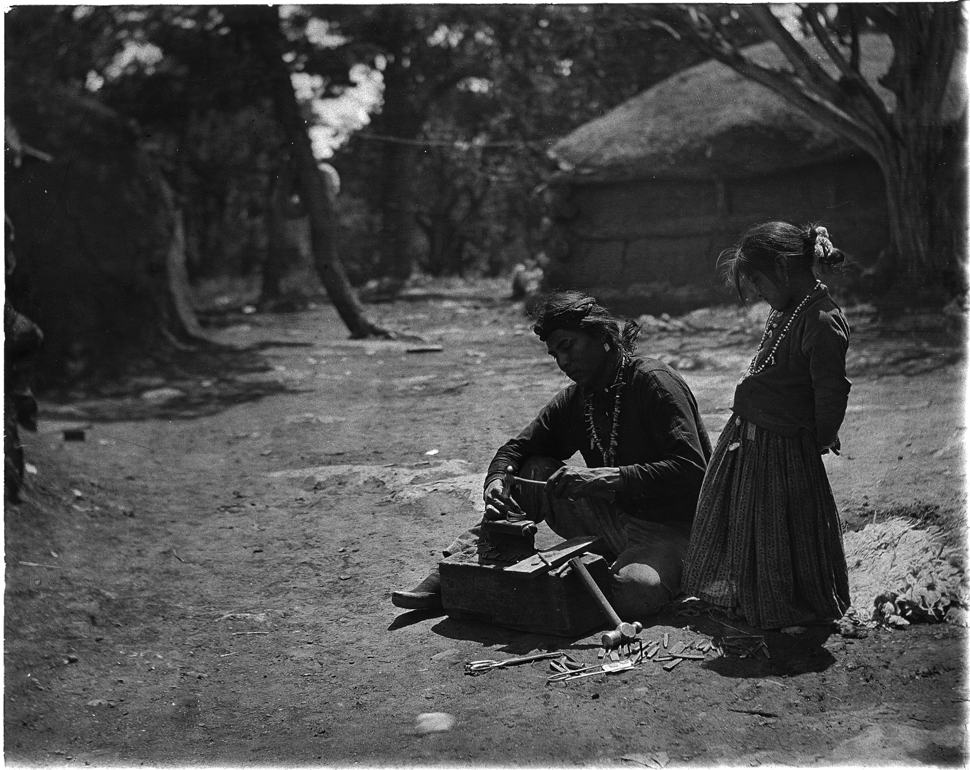 Black and white photo of a child standing next to a sitting adult hammering at an object. Behind and to their left are trees and a house-like structure. 