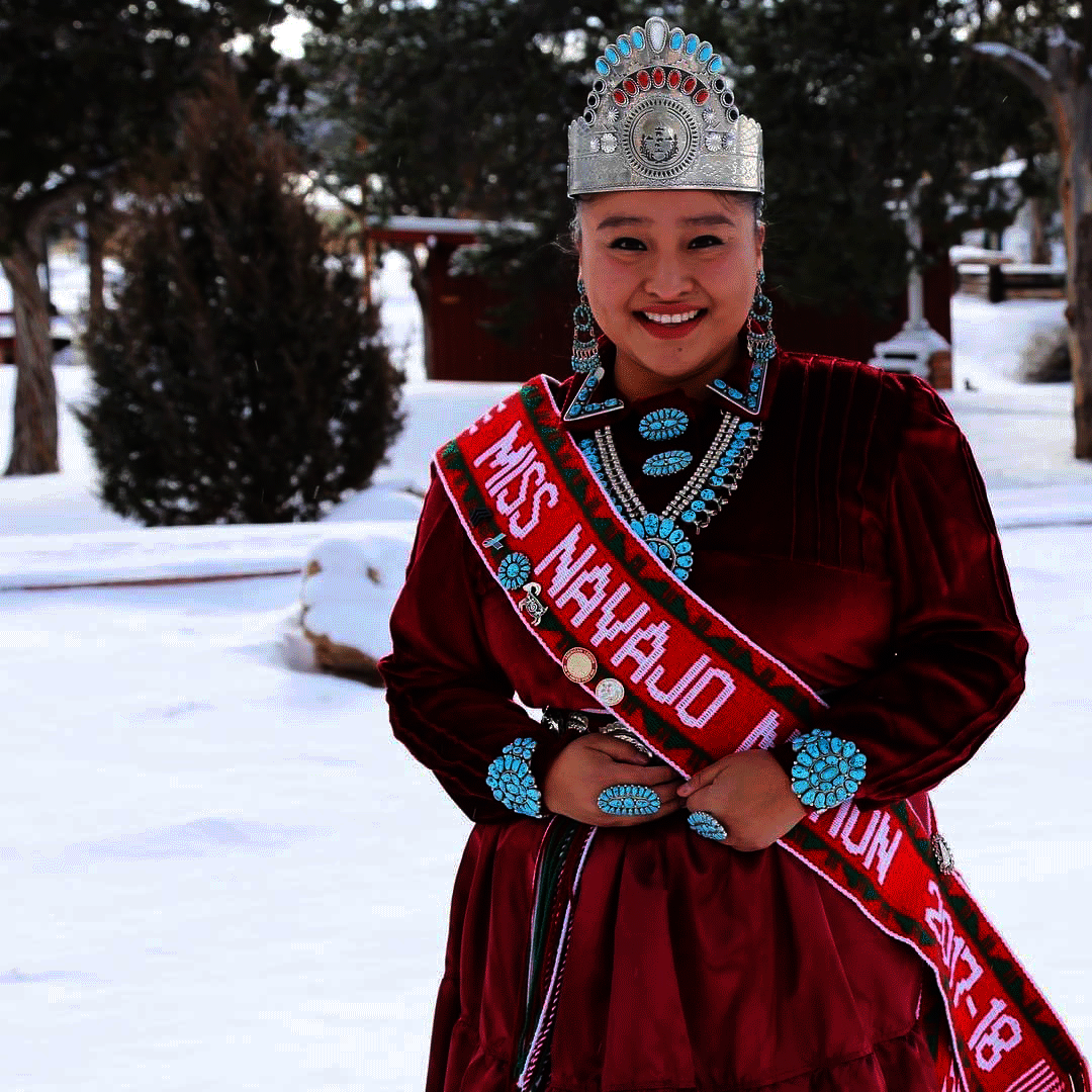 Photo of a person standing behind a snowy landscape, smiling wearing a silver crown with red and turquoise gems, and sash that reads, “Miss Navajo Nation 2017-18.” Their red velvet dress features turquoise detailing.