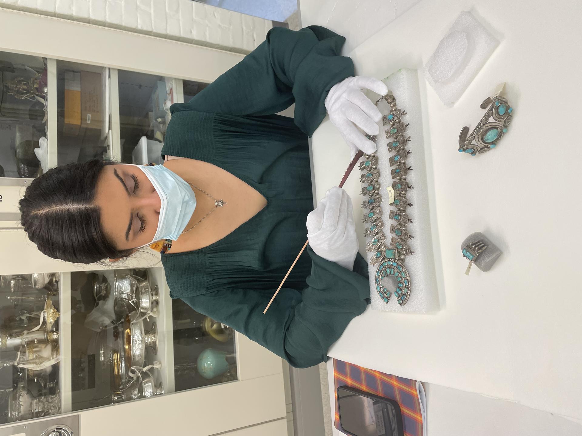 Picture of a person, wearing a mask and gloves, holding a long thin device to a silver and turquoise necklace laid in front of them on a foam rectangle.