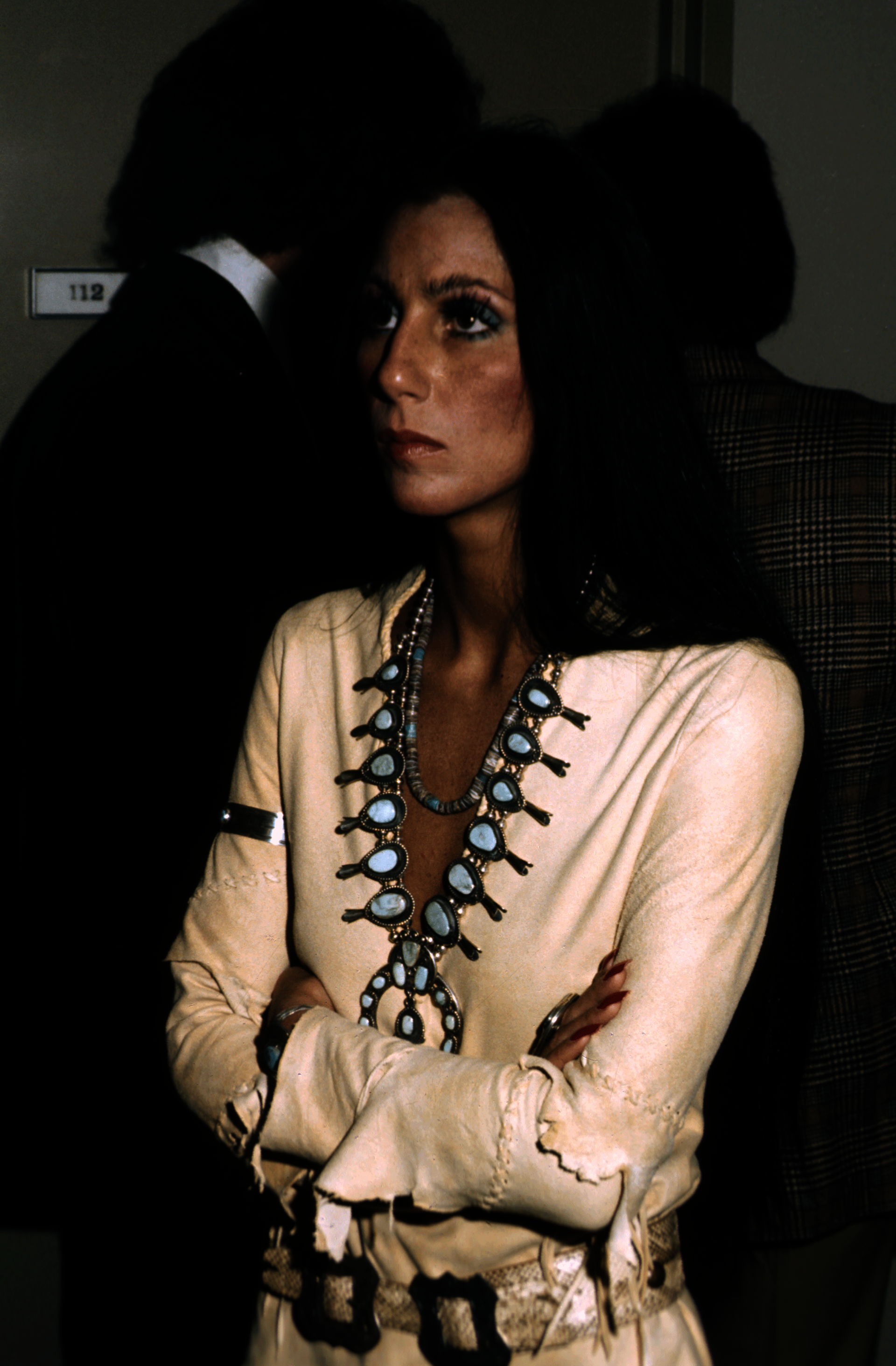 A woman with long black hair wearing white leather clothing and a silver and white pendant necklace. The person’s arms crossed, The woman is facing partly sideways with her arms crossed. 