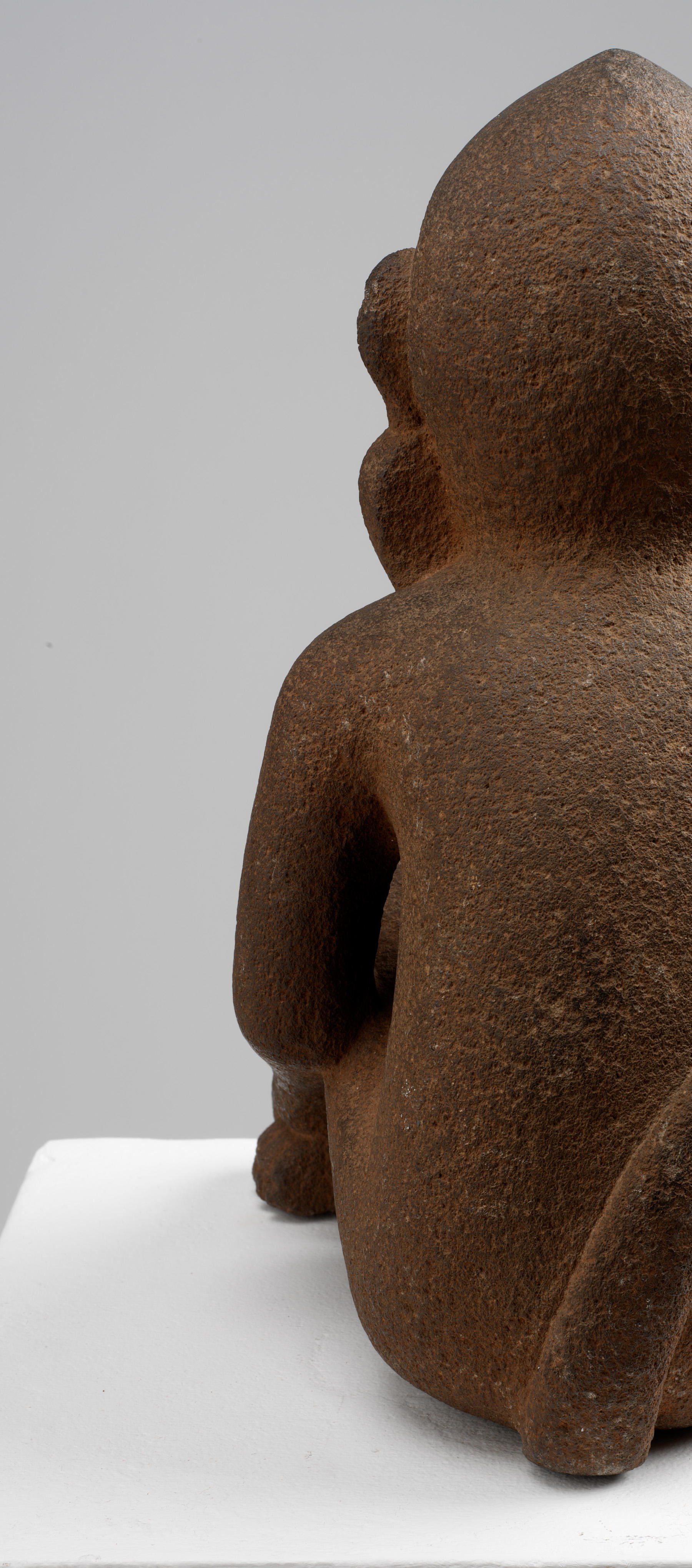 Cutoff back-view of the brown monkey sculpture’s left side. Its earrings are attached to the ear, and its tail goes upwards. The monkey’s texture is rough and porous.