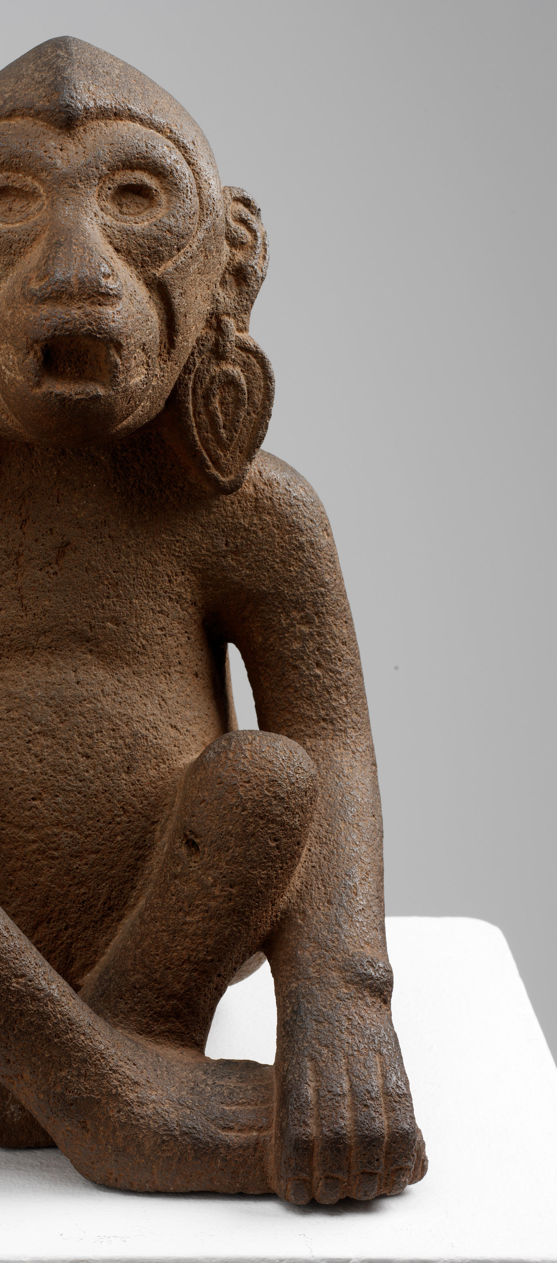 Cutoff view of the front right-side of a brown monkey sculpture sitting cross-legged with one hand gripping its foot. Its hands and toes are engraved to delineate toes and fingers.