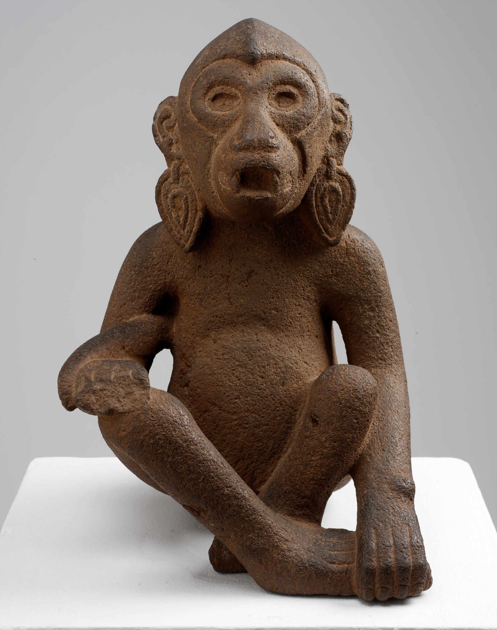 Front of a brown sculpture monkey wearing earrings. Its tail drapes over the left arm, and its right arm holds its left foot. Its eyes and mouth are open.