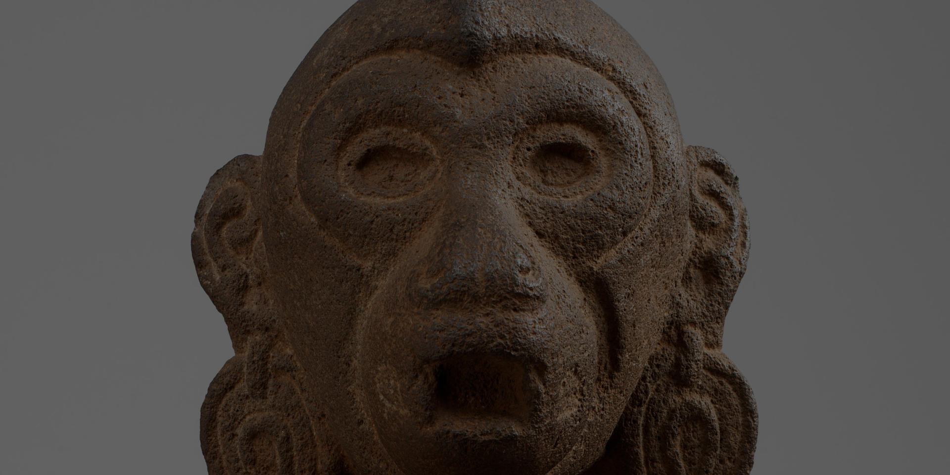 Detail of a dark brown, porous sculpture of a monkey head with a gray background.  The monkey has wide open eyes, large earrings, and an open mouth.