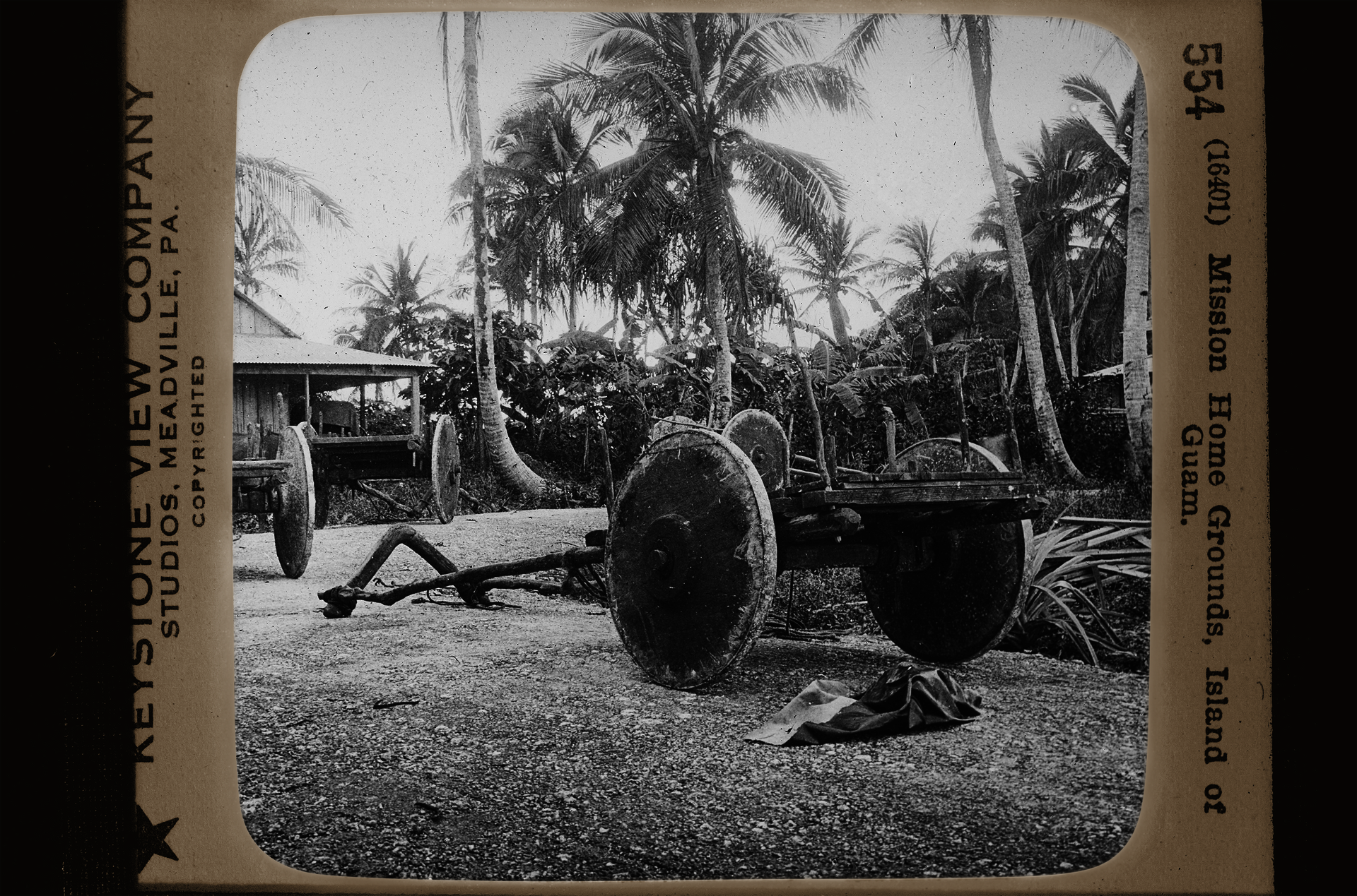 A black and white slide photograph of several 2-wheeled carts with large wooden wheels. A wooden structure with a porch sits in the left of the frame and large palm trees are in the background. Text on the slide frame reads 'Keystone View Company Studios, Meadville, PA. Copyrighted' and '554 (16401) Mission Home Grounds, Island of Guam.'