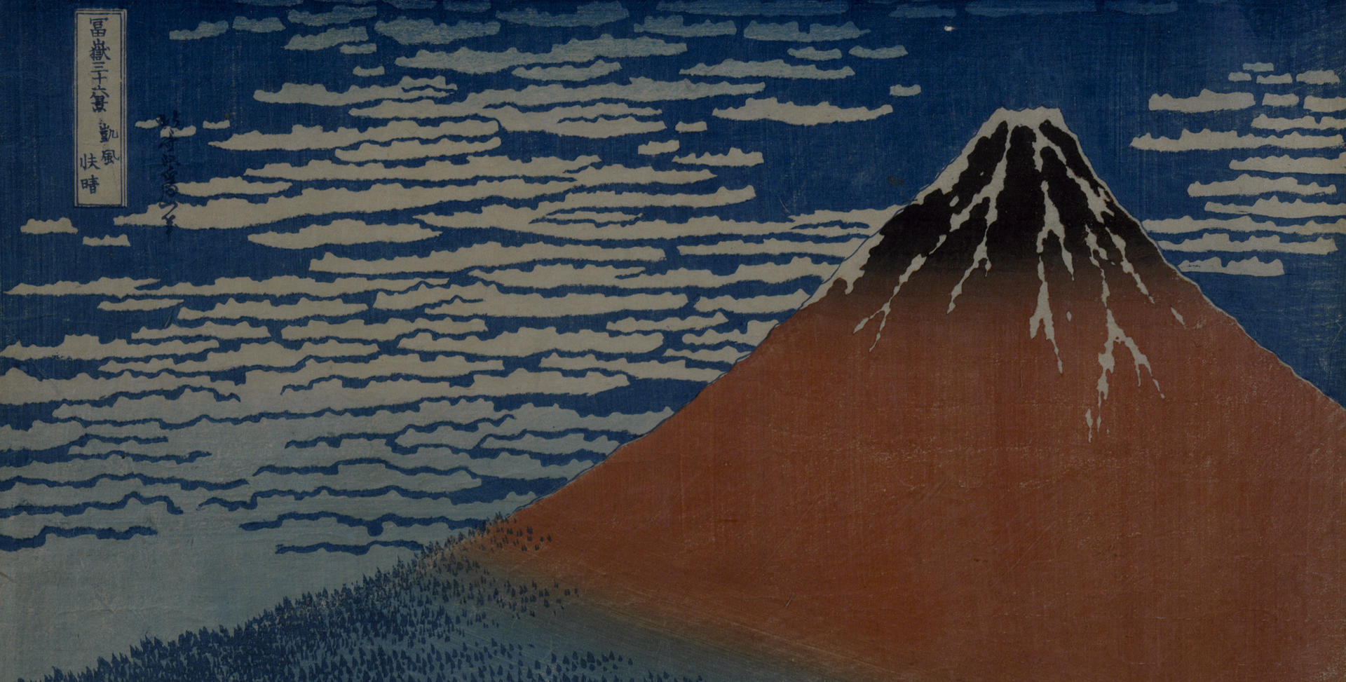 Woodblock print of a large mountain colored red and blue with white clouds and a blue sky behind it. Small trees sit at the base of the mountain. In the top left of the frame, Japanese text is in a white rectangle.