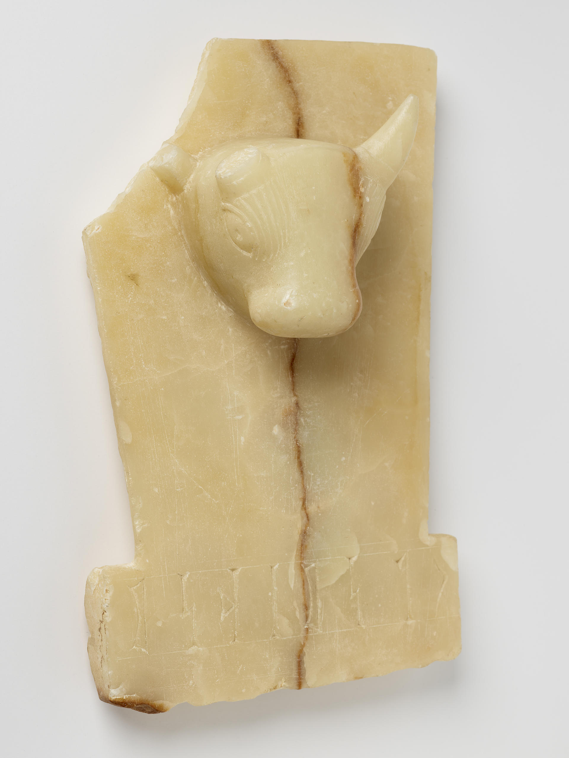 Cream rectangular stone with a sculpted bull’s head emerging from the upper center. Its upper left is broken off. A brown crack runs vertically down the center.