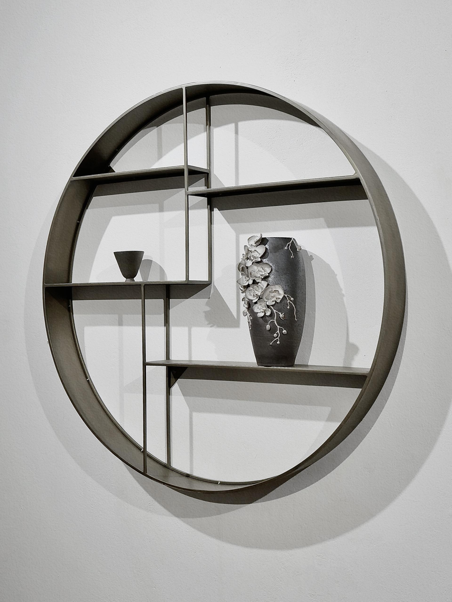 A dark brown Soda-fired ceramic vase with white hand-sculpted orchids and a small tea cup are displayed on a circular wall shelf.