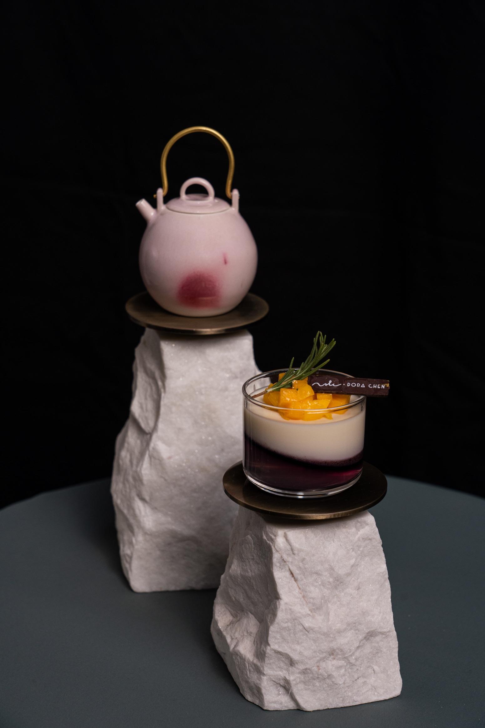 A pink glazed ceramic teapot with a brass handle and a panna cotta dessert in a small glass cup is displayed on two stone stands.