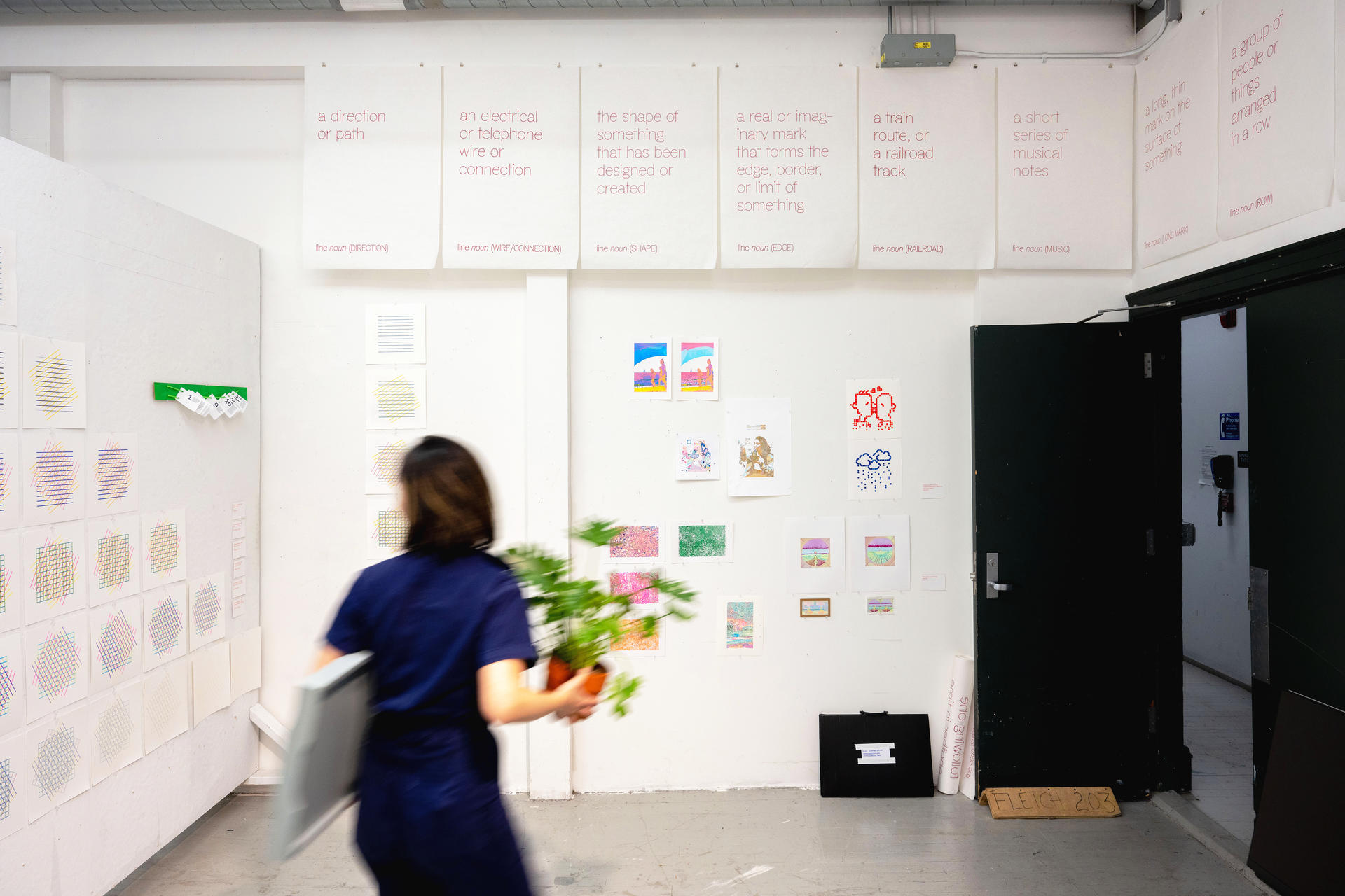 A room with a variety of small-scale works pinned to white walls. A person in motion blur walks in front of it, carrying a plant.
