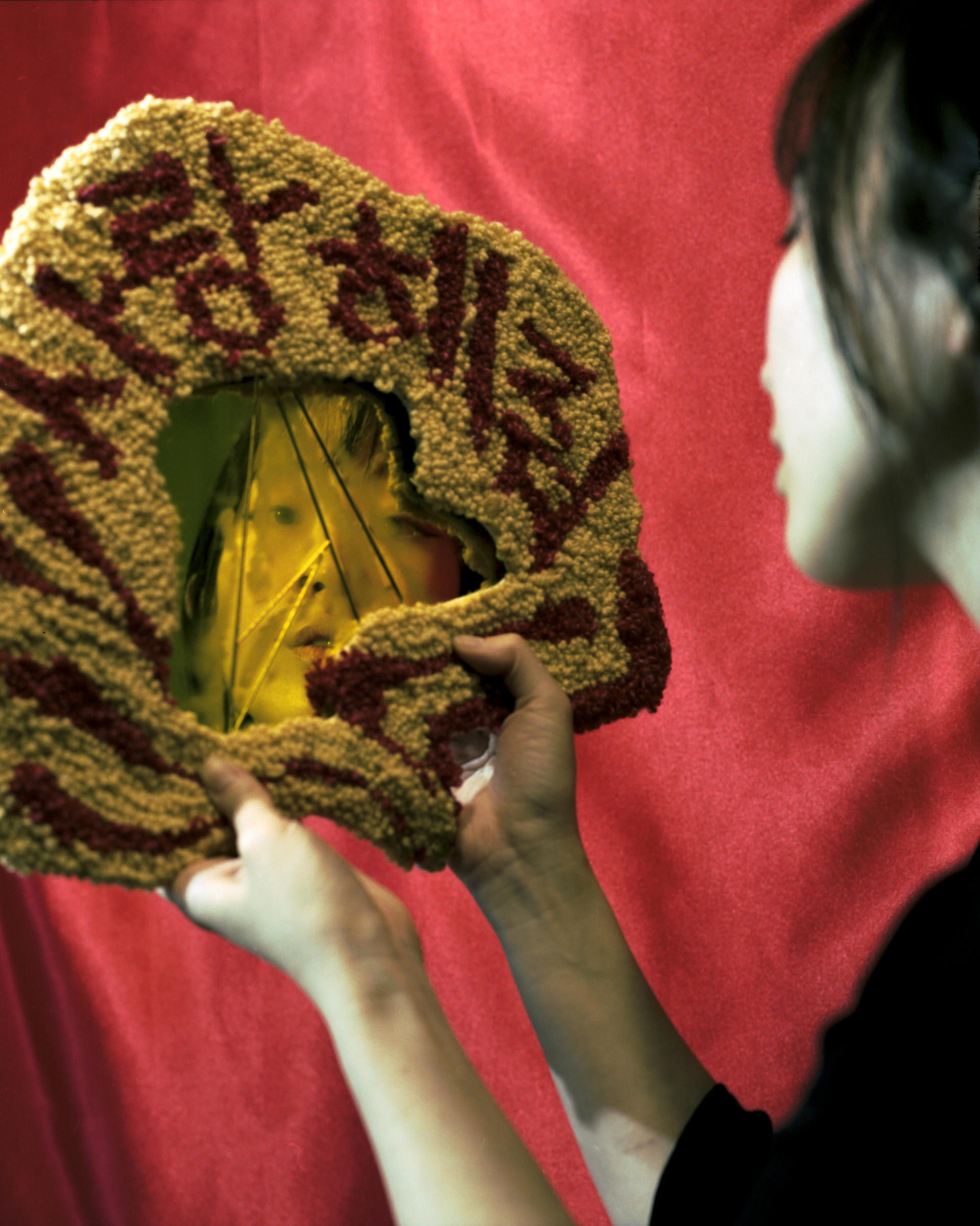An asian woman looking into a fractured mirror covered with a tufted frame displaying the words "Love Me" in Korean.