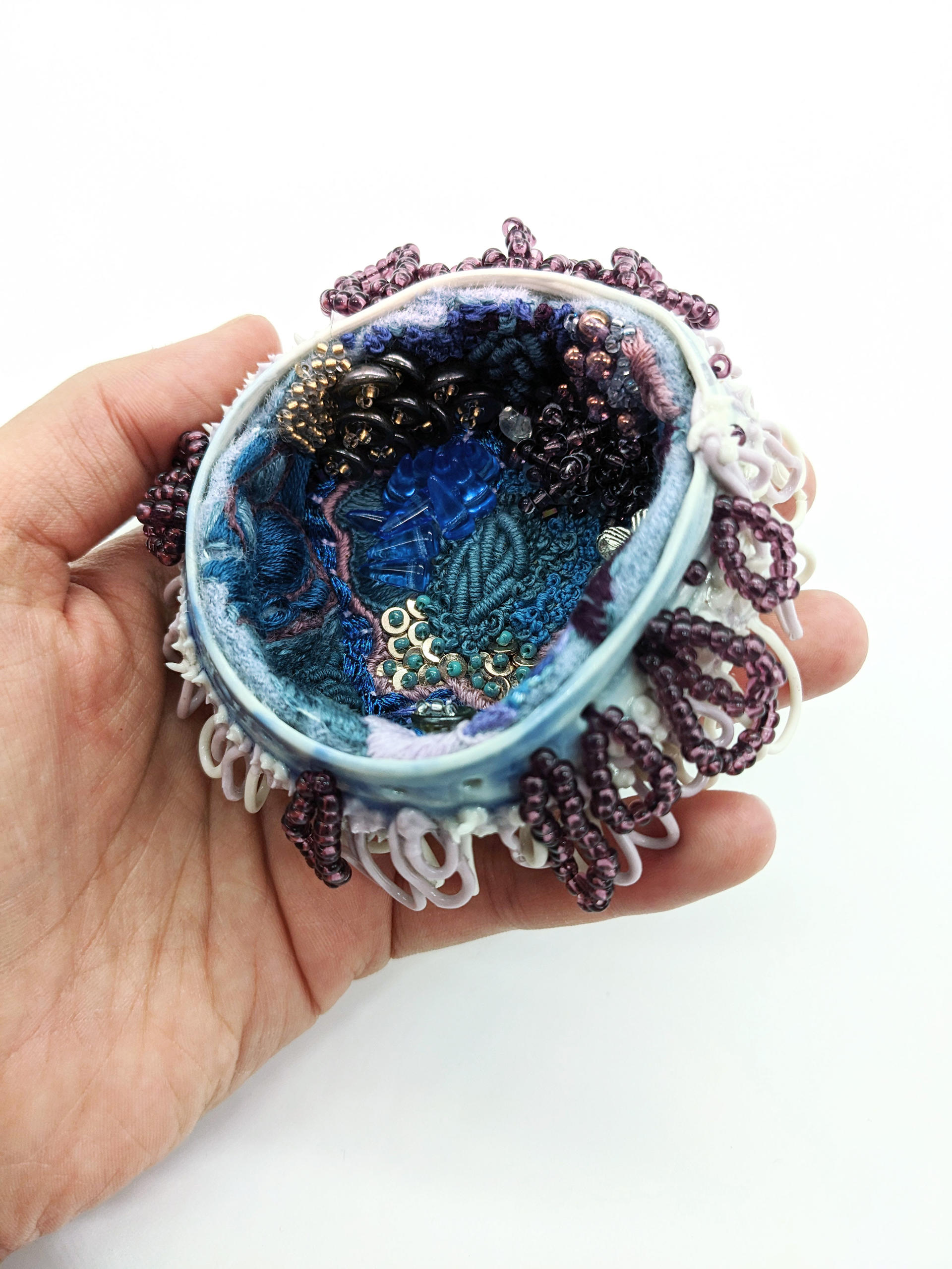 hand holding small porcelain vessel lined with beads and embroidery
