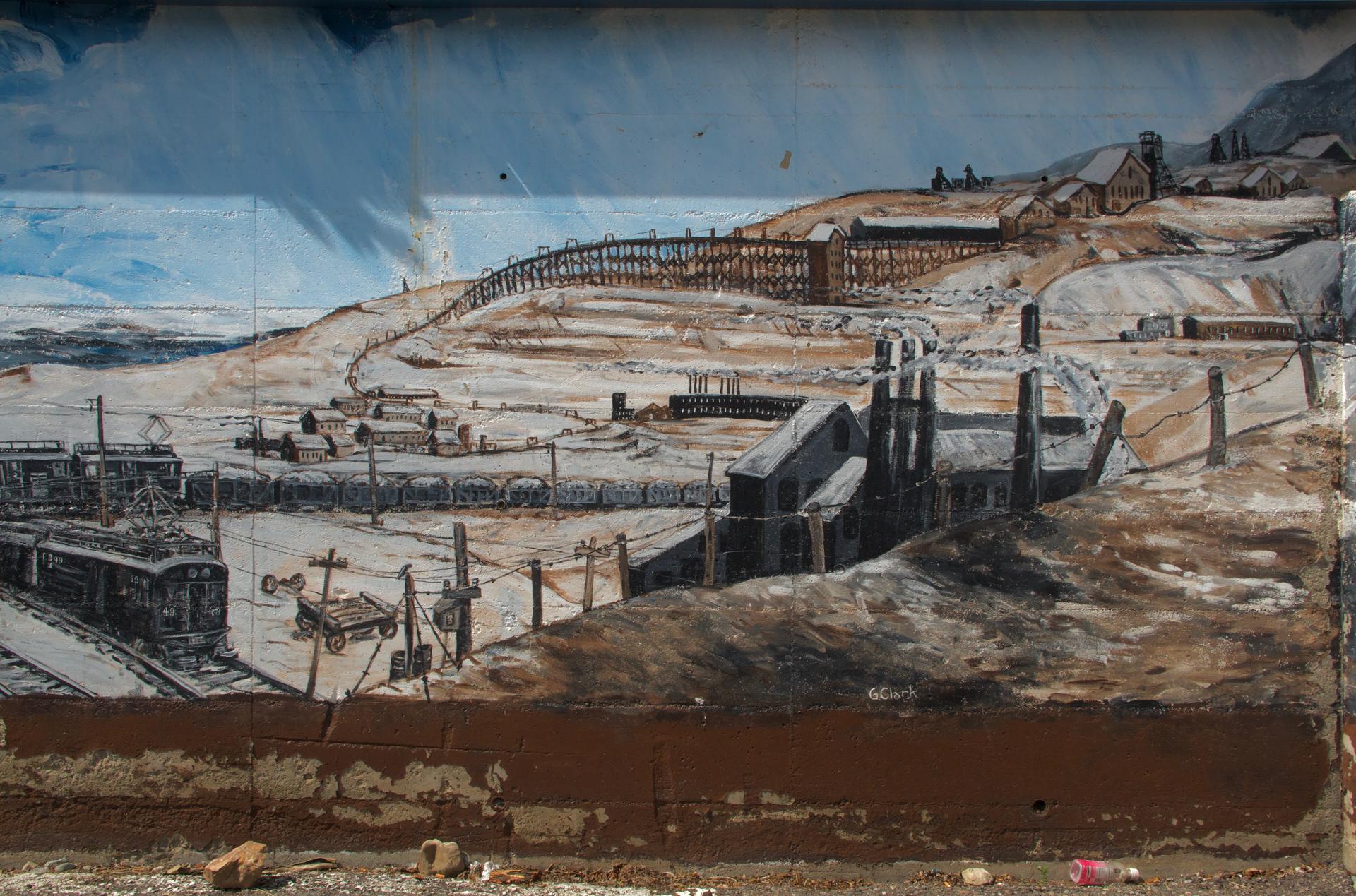Photograph of landscape mural depicting the settler copper industry in Butte, Montana in the early twentieth century. A shadow of a plant is visible against the mural’s blue-painted sky. In front of the mural, pieces of trash lie strewn about.