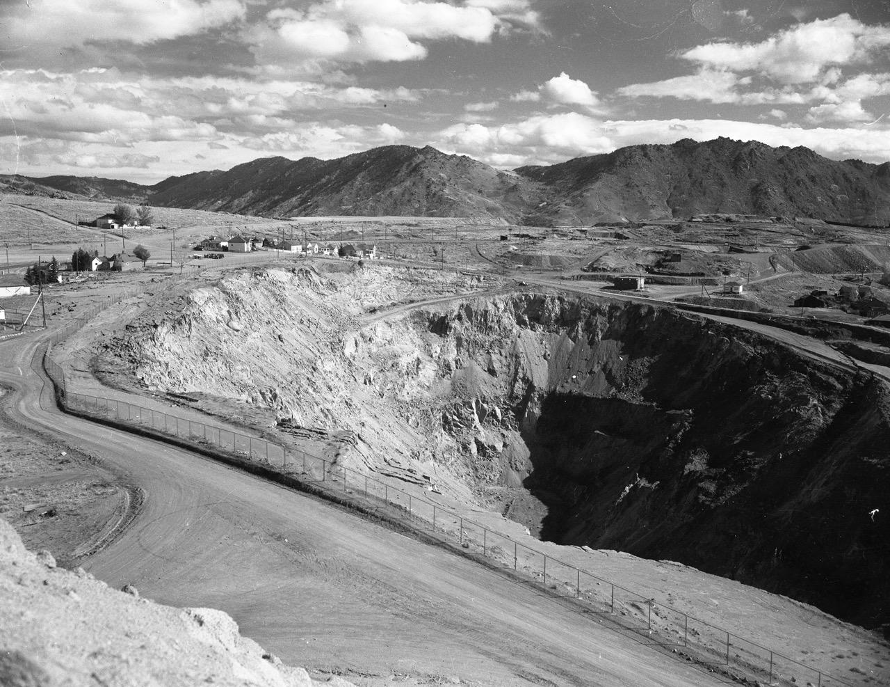 An open-pit copper mine with a caved-in wall gapes open in the foreground. A shadow blankets the southern rim of the pit. The heavily mined ground beyond the pit slopes up to meet the Continental Divide, from which water flows in two directions. Here is the source of Silver Bow Creek, draining into the Pacific Ocean.