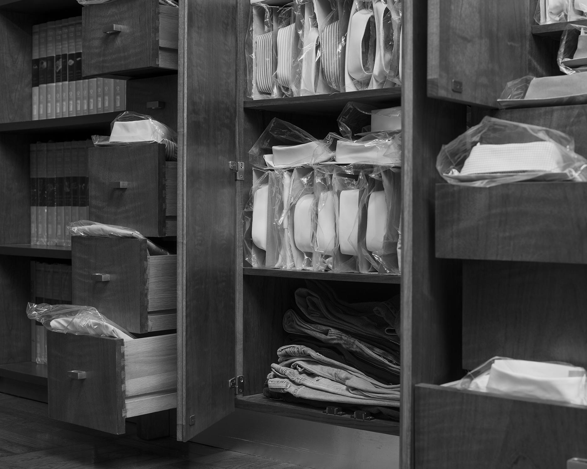 Black and white pigment print of folded shirts emerging from cabinets and drawers. 