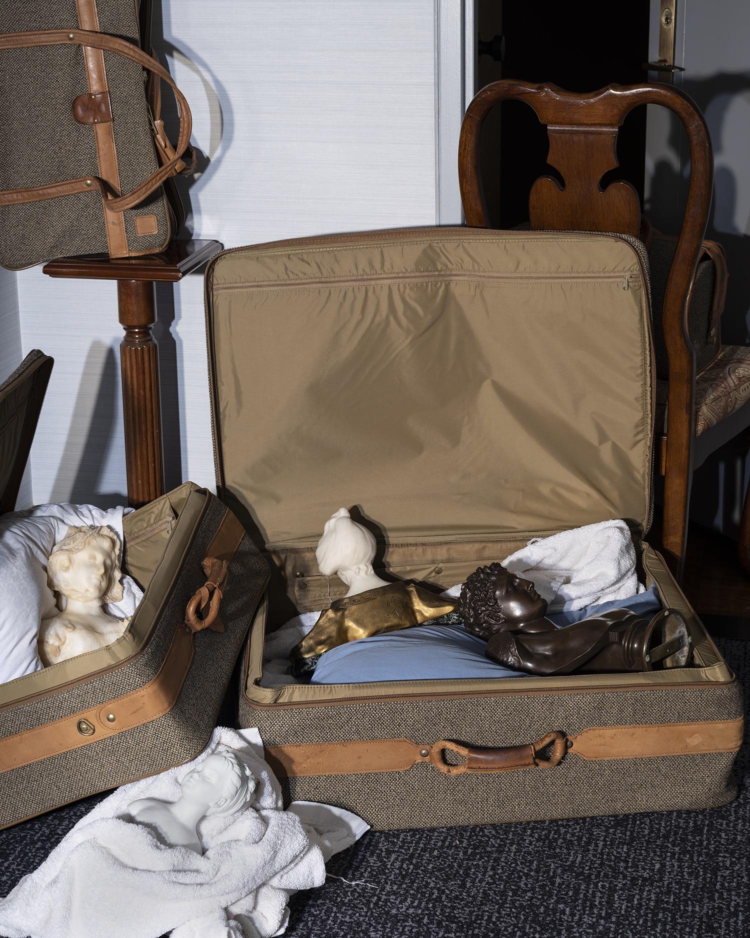 Color pigment print of busts in and around open suitcases. 