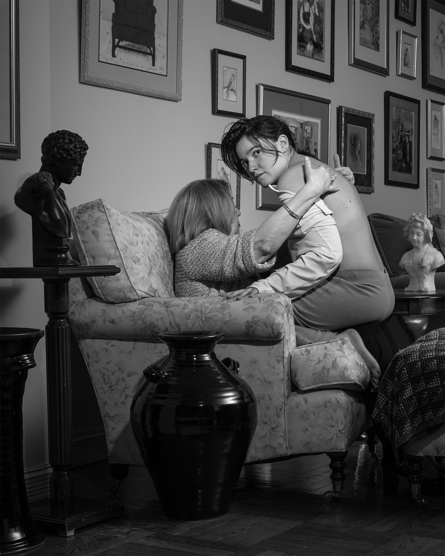 Black and white pigment print of young adult sitting on older adult's lap surrounded by framed images, vases and busts.