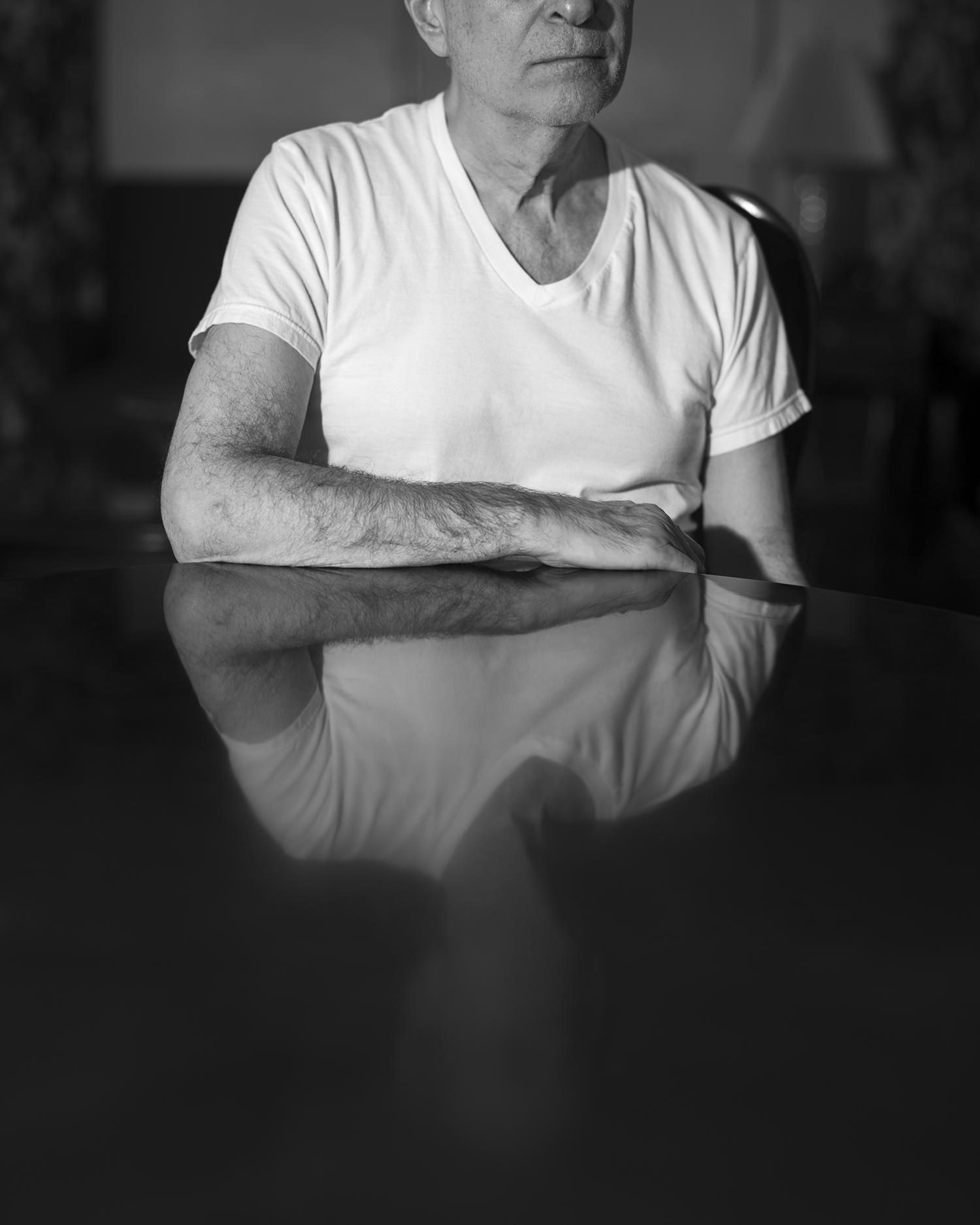Black and white pigment print of man sitting with his obscured image reflected in dining room table.