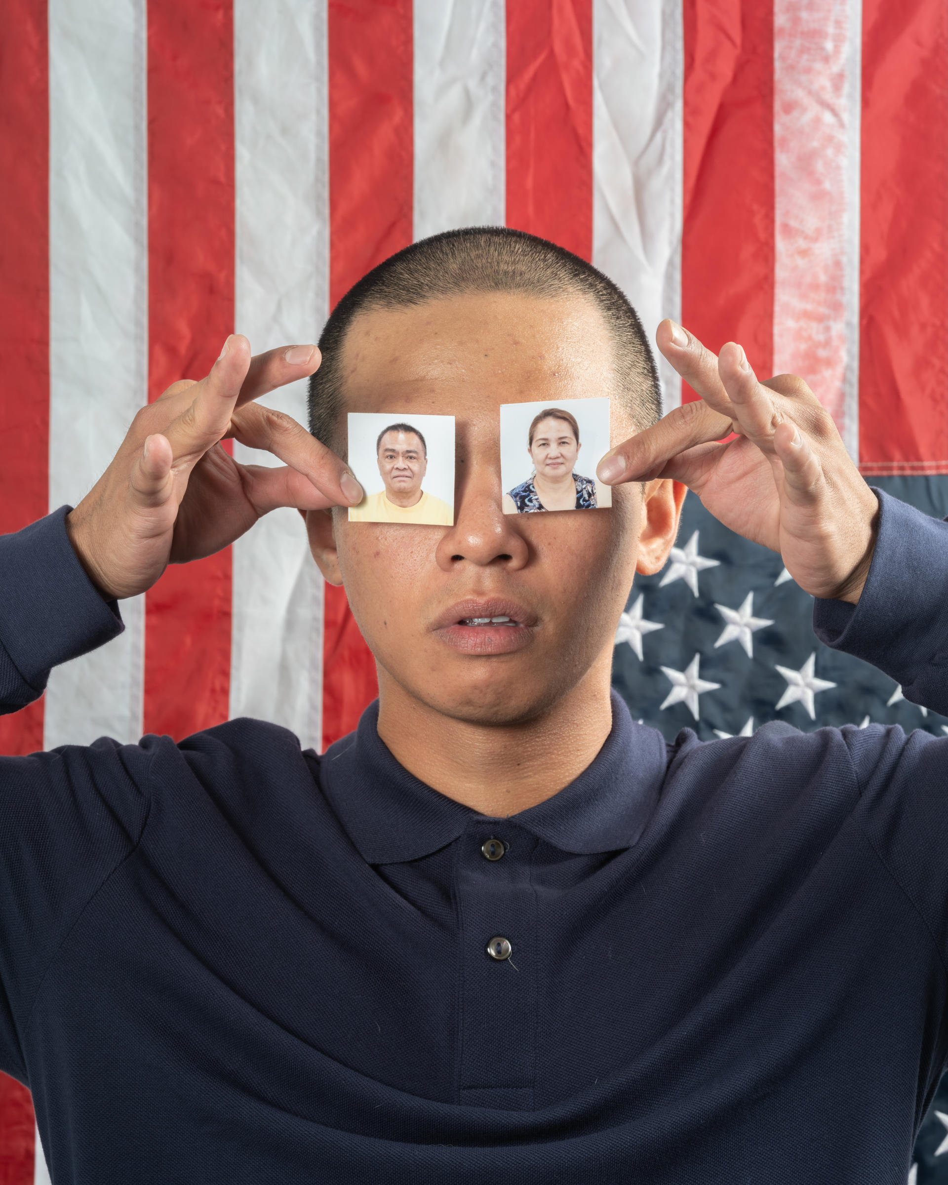 A person holds up two passport photos, one in each hand, covering their eyes with it against an American Flag