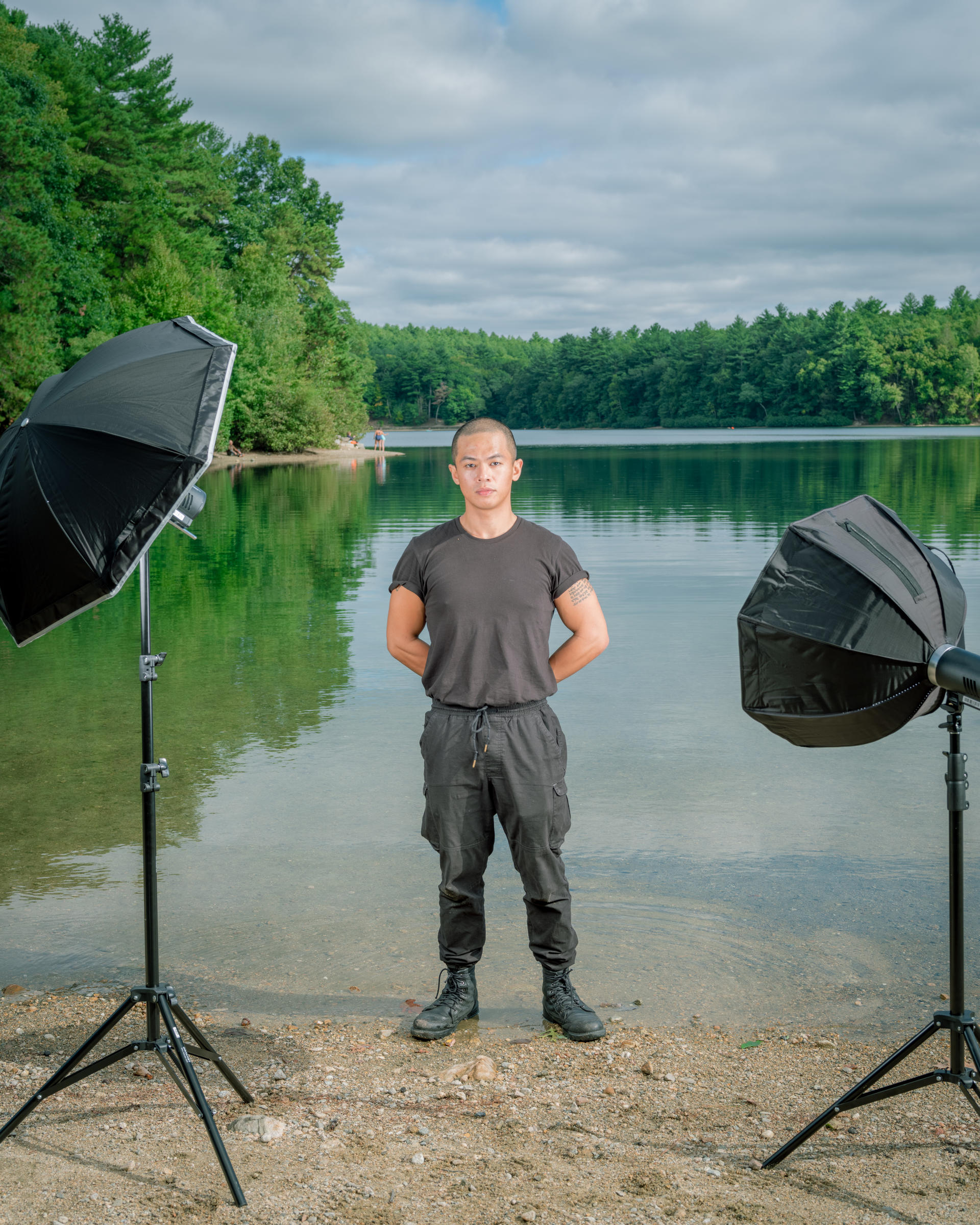 A man wearing all black standings shoulder-width apart at the shore of lake. Two flash strobes frame him.