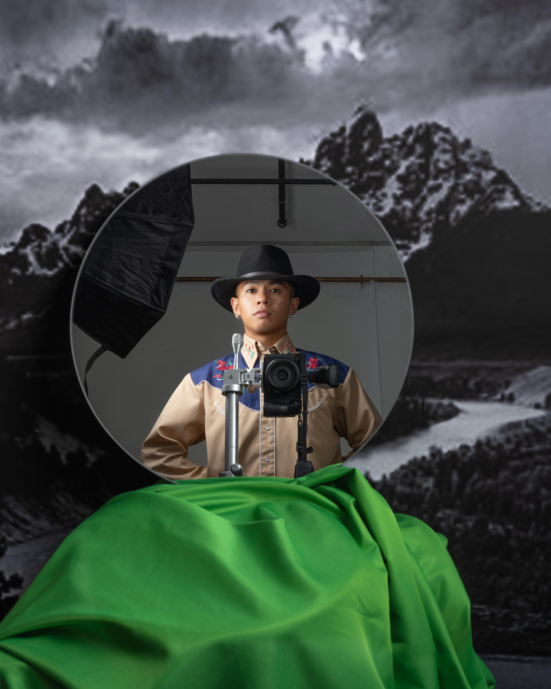 A mirror shows a cowboy standing behind a mounted camera. The mirror is propped against a print of an Ansel Adams landscape.