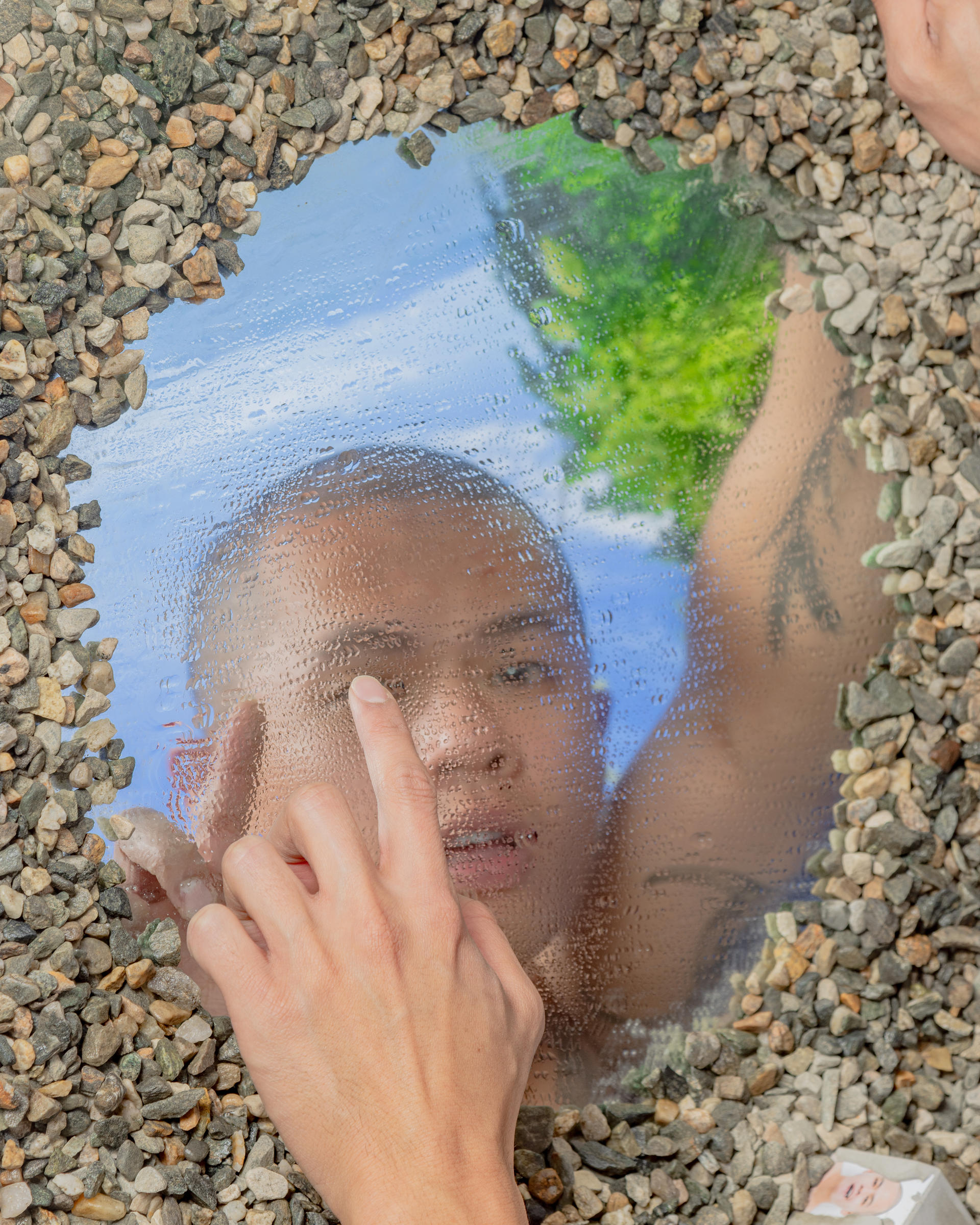 A man is touching a circular pool showing his reflection, the pool is surrounded by gravel.