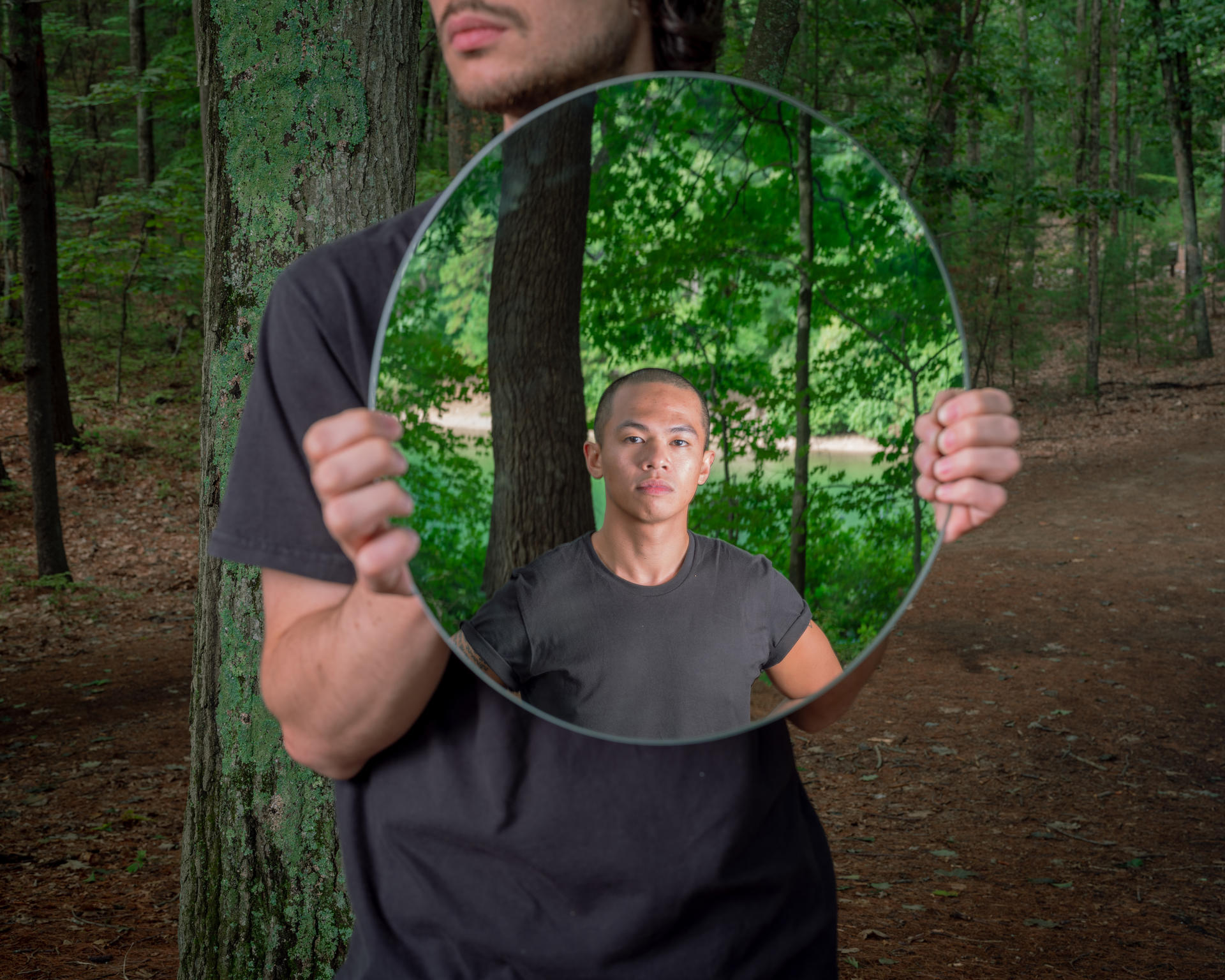 A man in a black shirt holds up a mirror showing another man wearing black. They are in a wooded area.