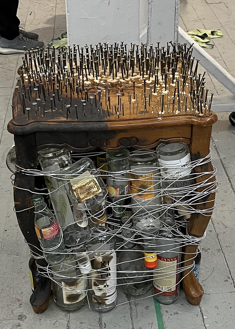 Side table wrapped with with wire and empty alcohol bottles. Hold of the table is burnt and rusted nails cover the top of the table except for a flower printed onto the table