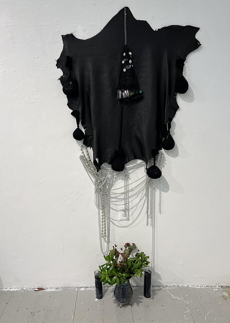 Black leather with several sacks sewn around the edges. A weaving including a glass hot sauce bottle hangs overlapping. Beneath on the wall dangles silver Mardi gras beads. Beneath on the floor are two black prayer candles, a silver vase containing spiritual herbs.