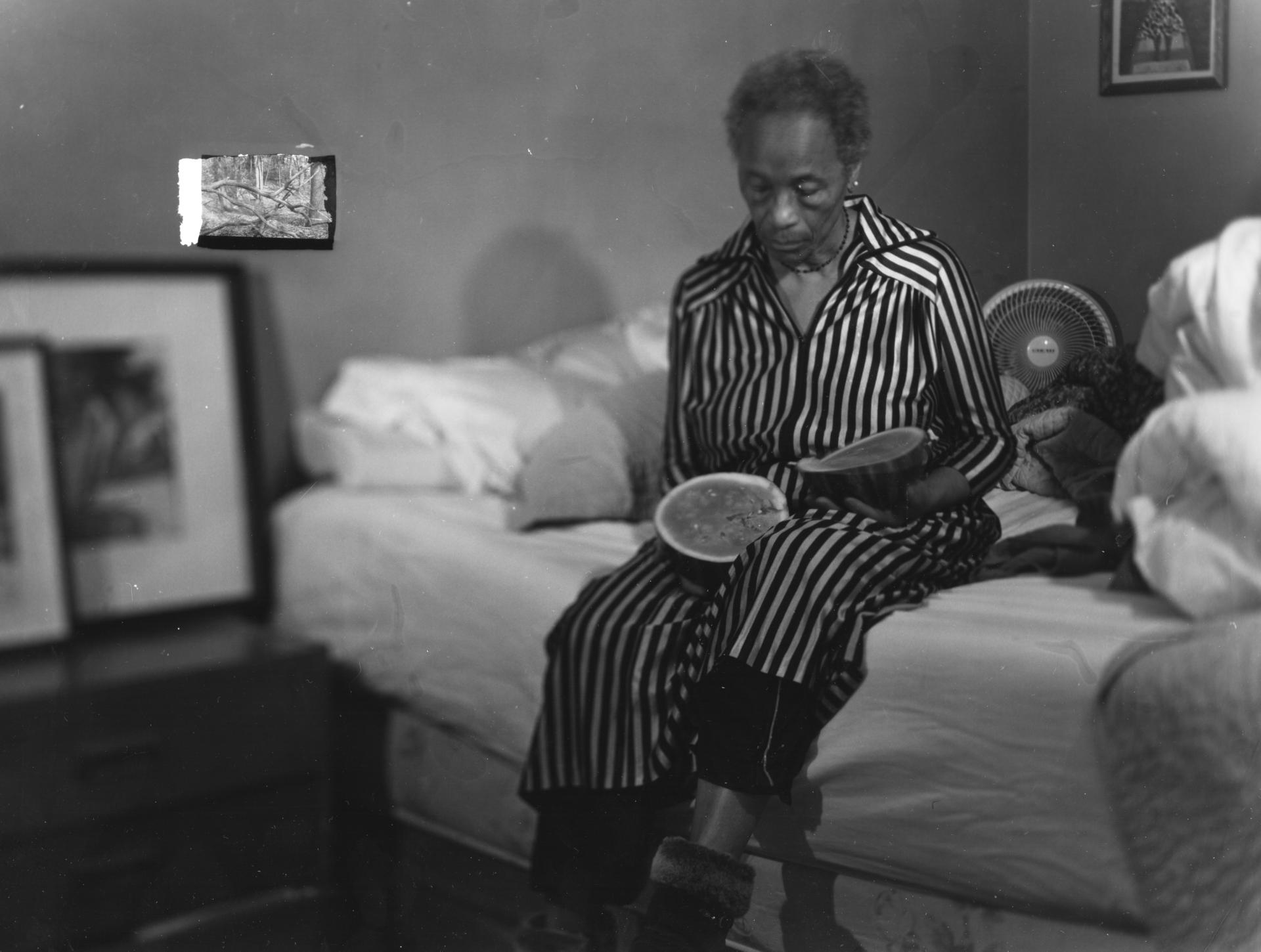 woman sits on bed curiously holding a fruit. in the corner, a glitch appears, a small inlaid image of a forest.