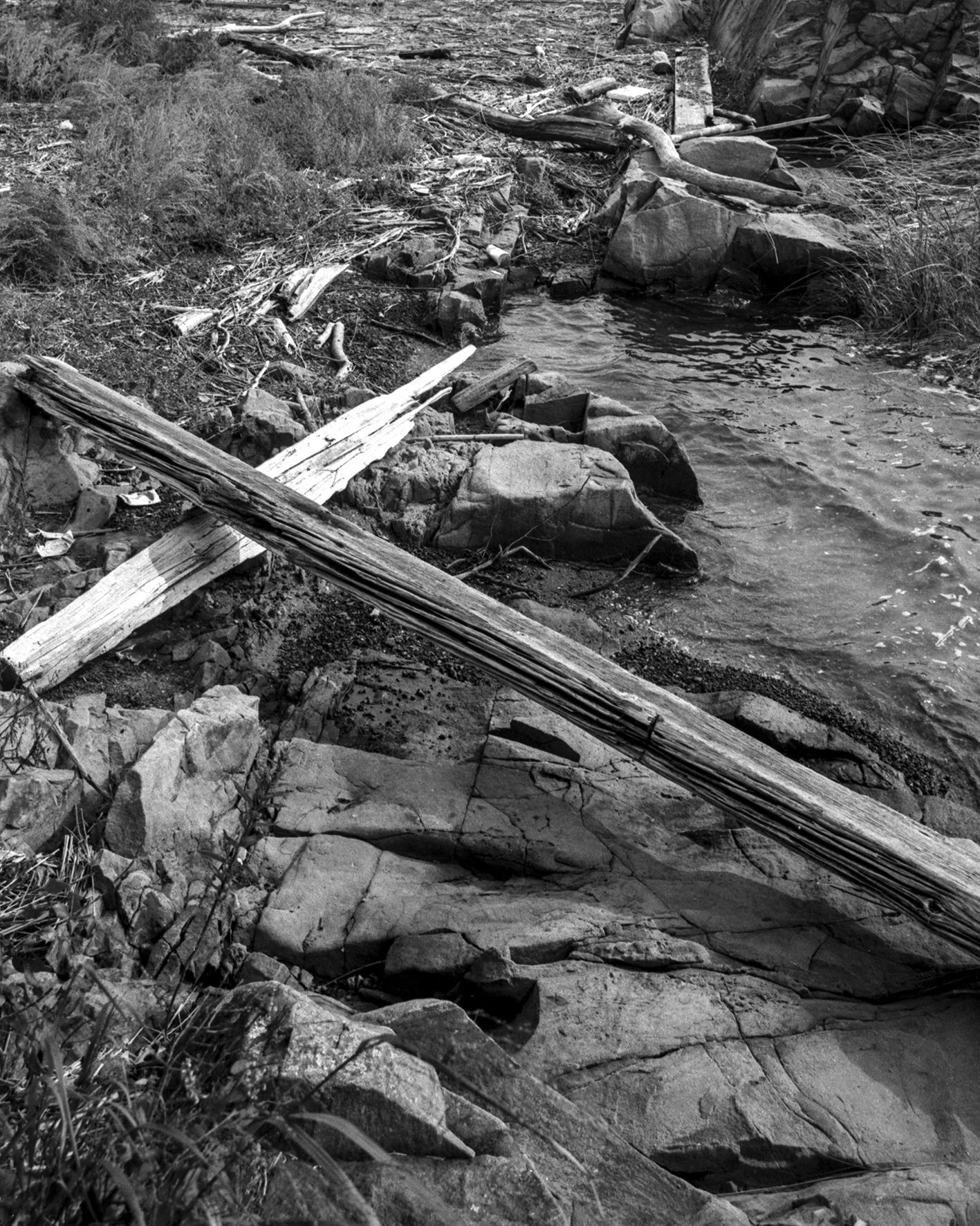 a black and white photograph of two long planks of driftwood in the shape of a cross, washed up on the rocky shores of a bay. 