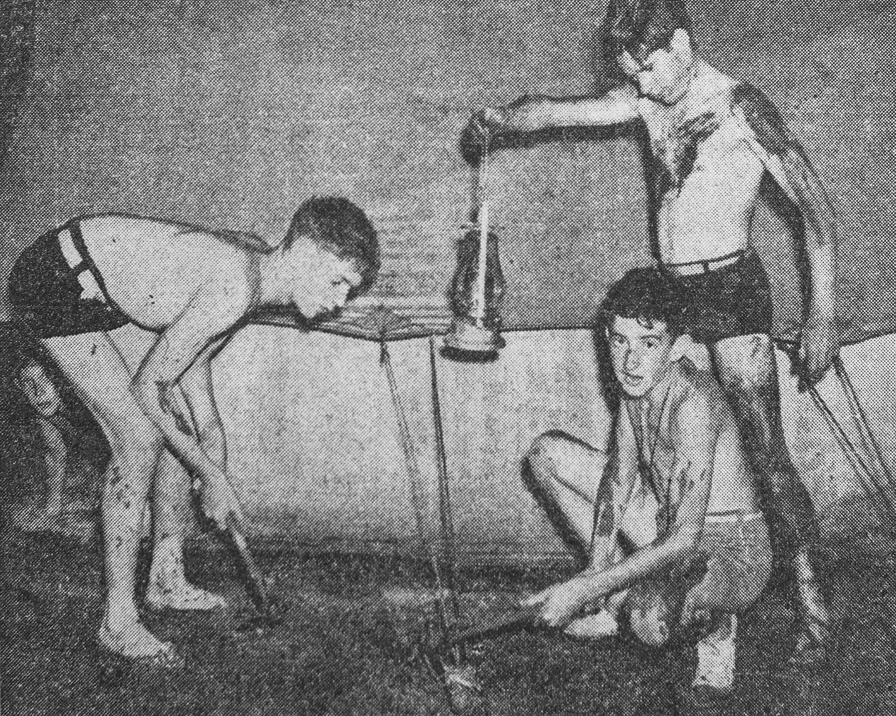 A black and white photo of three light skinned adolescent boys gathered around the side of a tent trying to repair it, it is raining and muddy in the middle of the night.