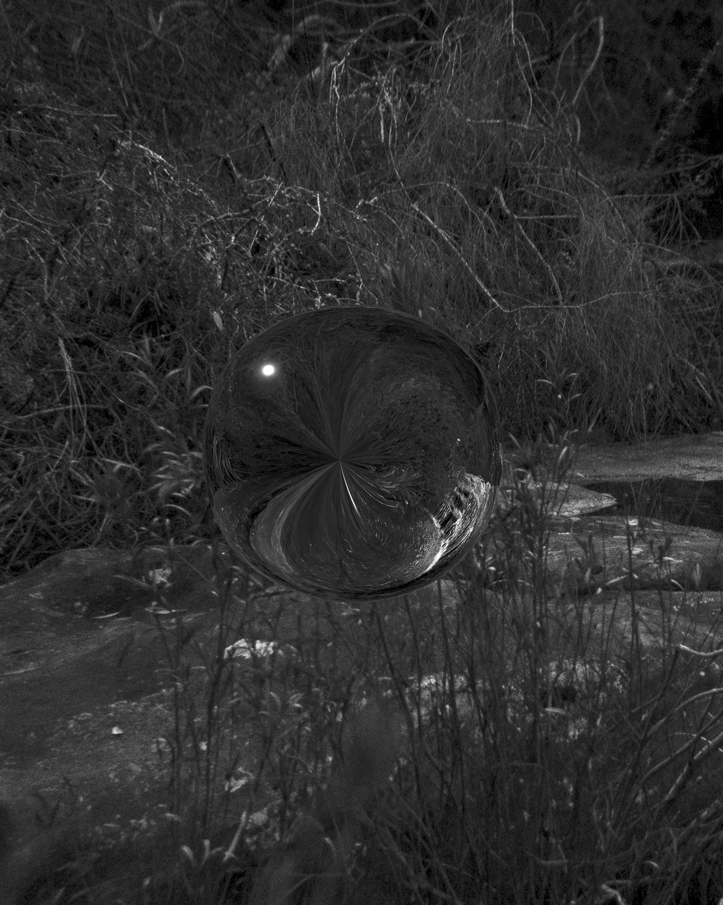 A black and white photograph of a reflective orb, floating in mid air, at night, amongst bushy trees and a water creek underneath.