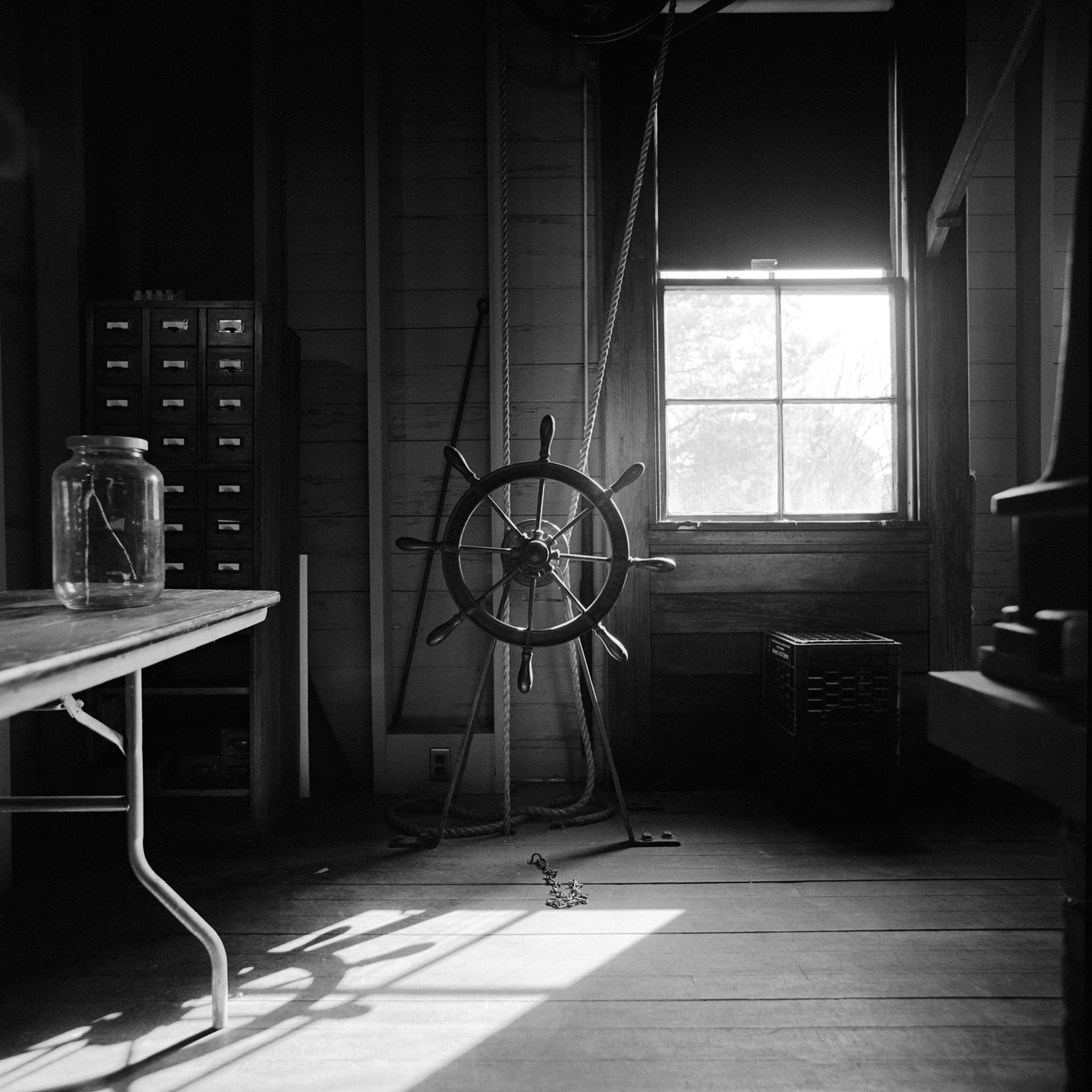 A black and white photograph inside an old looking observatory picturing a wooden ship wheel with ropes attached around it that extend up towards the ceiling.