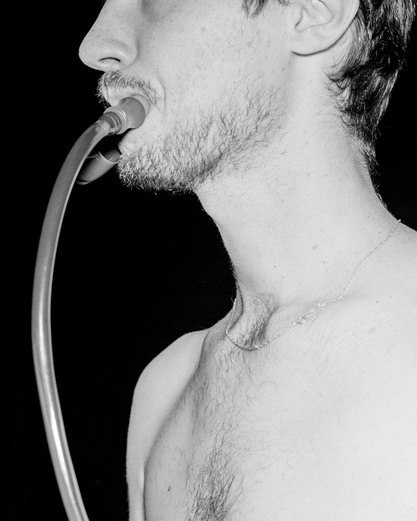 A black and white photograph of the chest, neck and jaw of a light-skinned man drinking from the hose of a hydration flask.