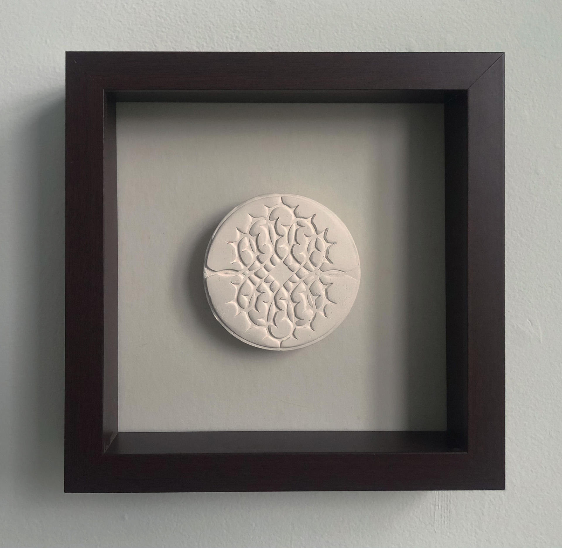 A pale beige ceramic disc with a thorn motif framed in a mahogany shadow box.