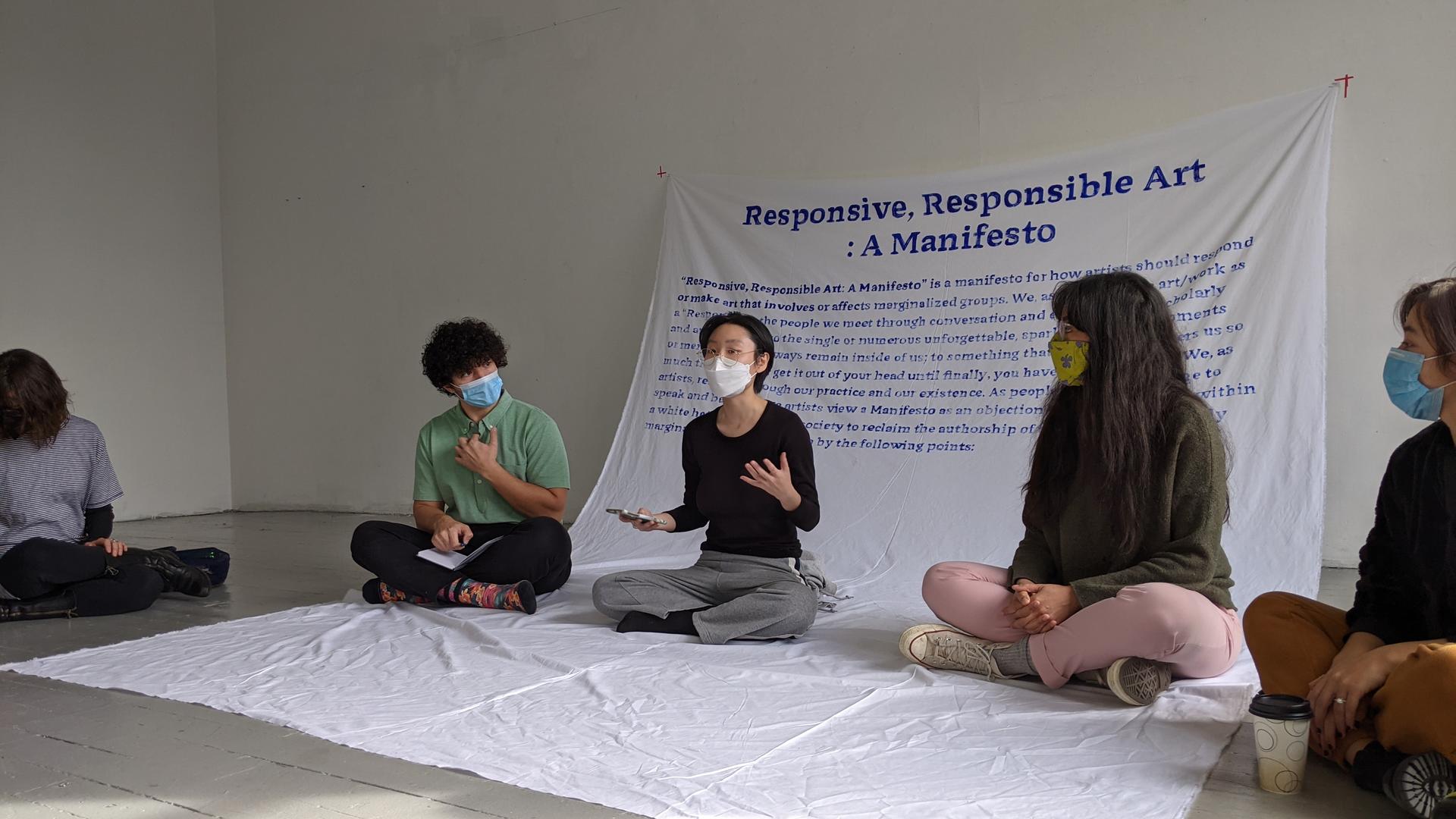 Artist reading “Responsive, Responsible Art: A Manifesto” on her phone while seated in a circle with four people on a large white fabric cascading down from the wall, featuring a handwritten paragraph from the manifesto in vivid blue paint.