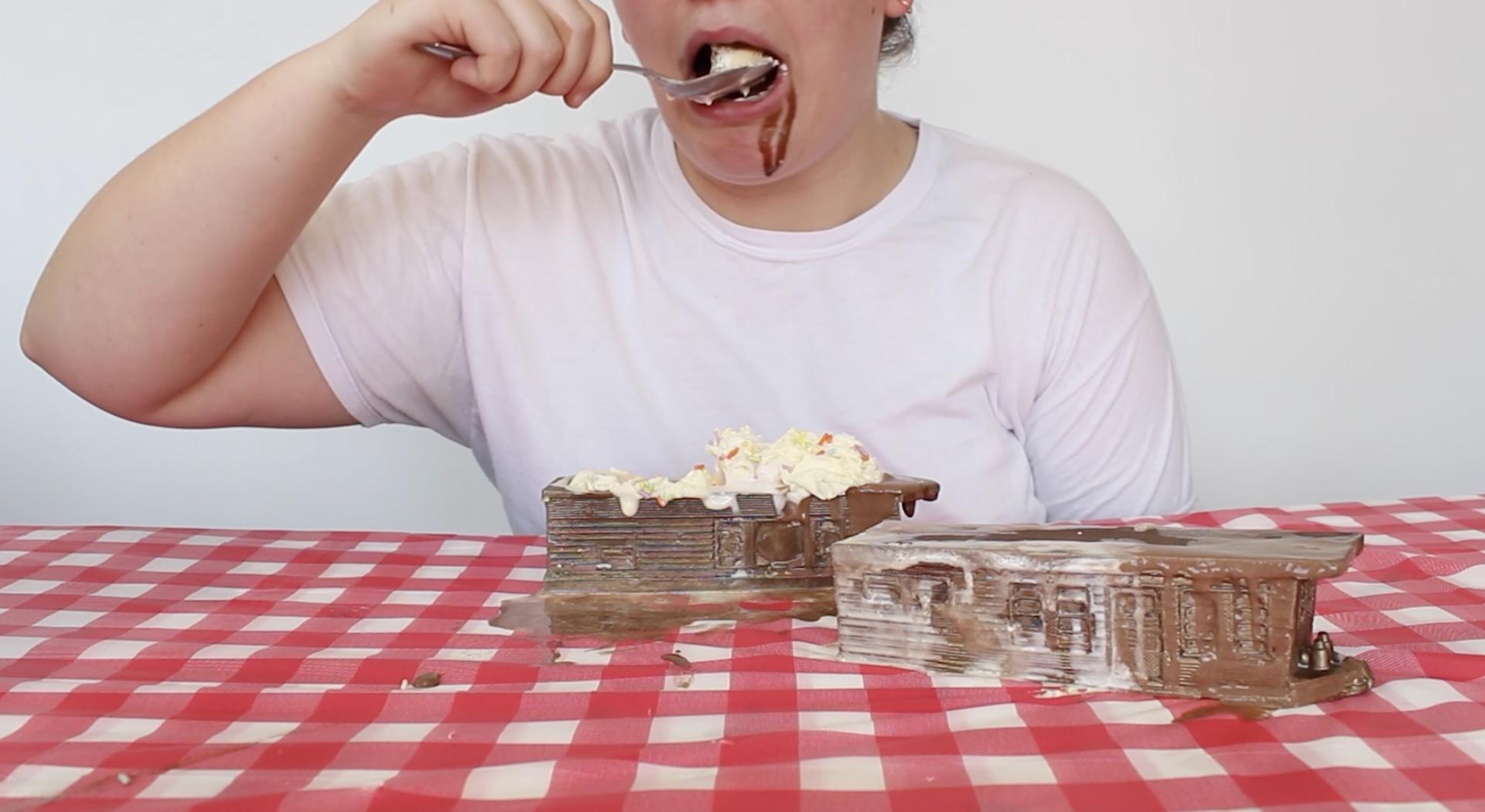 A white person sits at a picnic table behind two miniature bronze trailer homes. The trailers are vessels containing ice cream sundaes. The person inserts a large spoonful of vanilla ice cream in their mouth