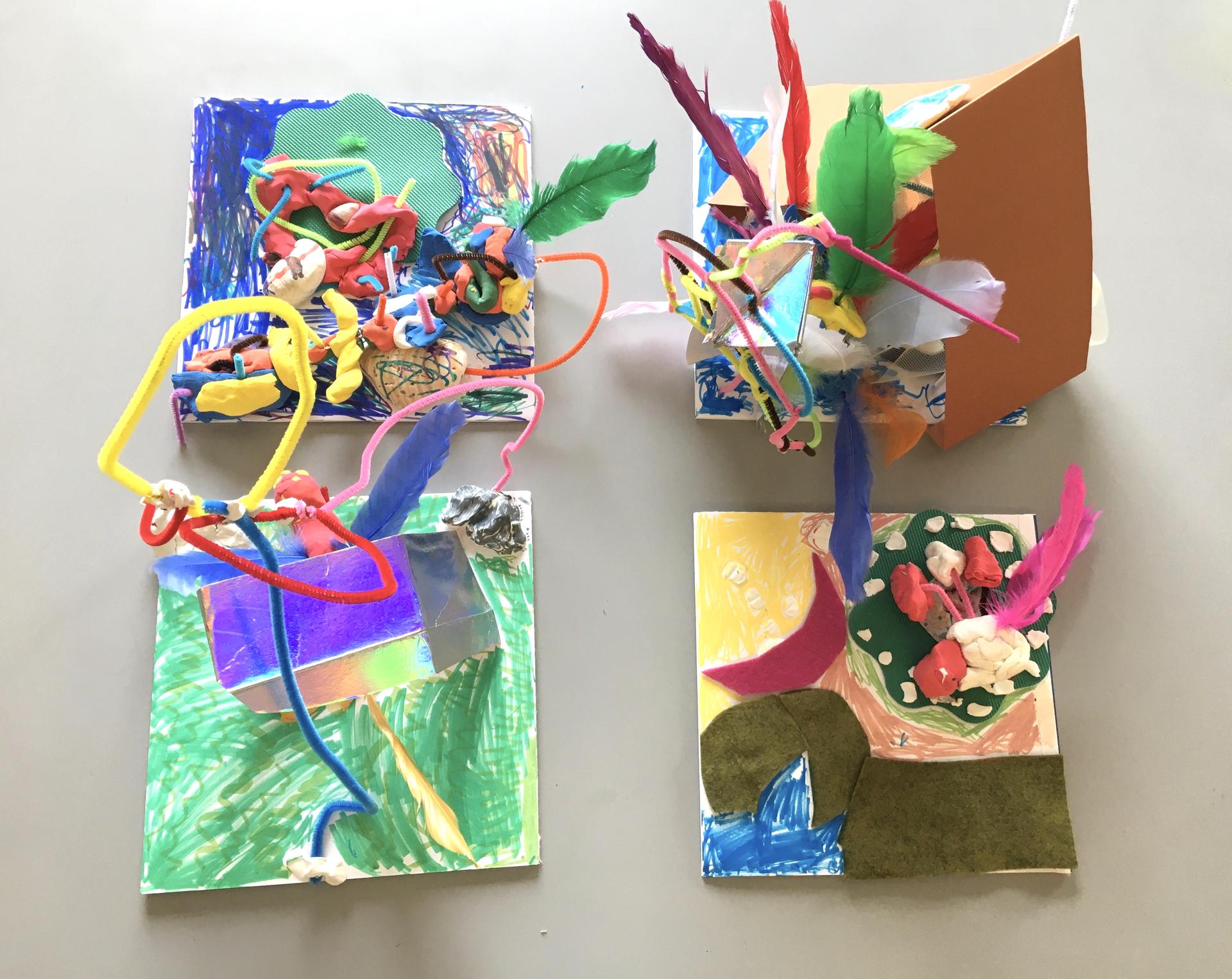 Four "habitats" built on square pieces of foam core, colorful air dry clay, pipe cleaners, shells, feathers, repurposed fabrics and paper, marker. Each habitat is constructed and colored in its own way, with some elements extending upwards towards the camera. 