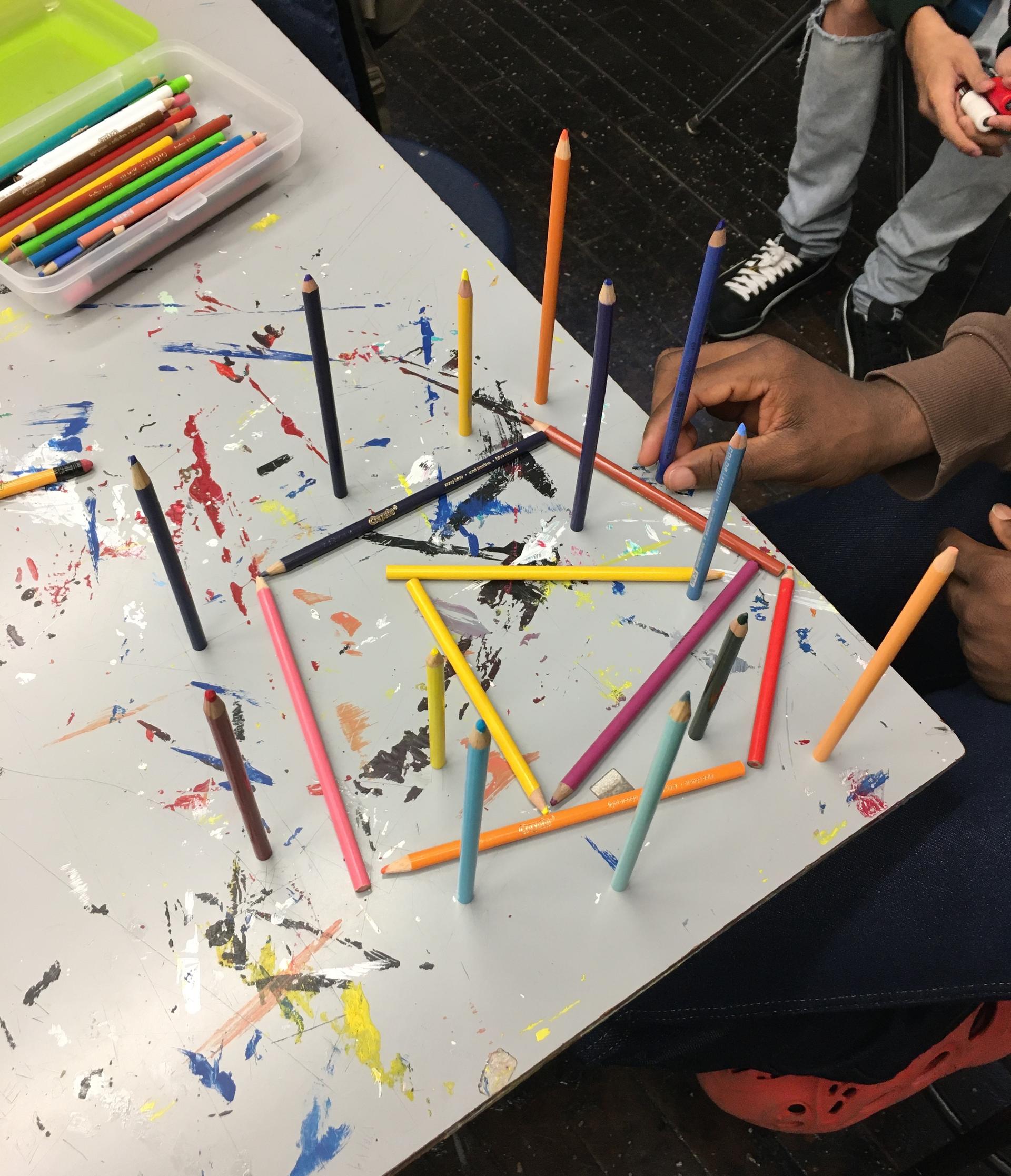 A student’s hand placing one of 13 colored pencils that are standing on their ends, pointing vertically upwards from a paint-splotched table. The vertical pencils surround 3 colored pencils lying flat on the table in the shape of a triangle, inside a pentagon made of 5 colored pencils. Another student’s legs and hand can be seen in the corner of the image.