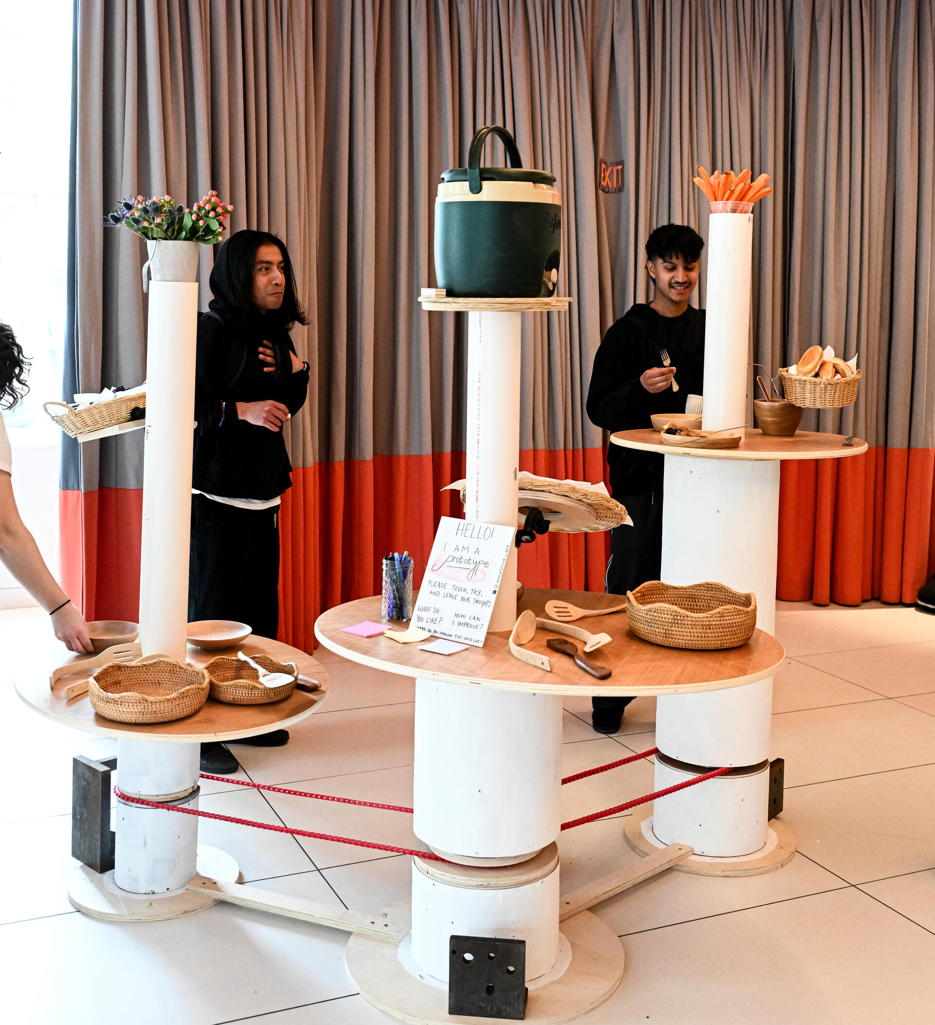 Three white columns with ovular tabletops and one tall cylindrical pipe coming out from each surface. The three tables are connected by a red mechanical belt which turns them. 