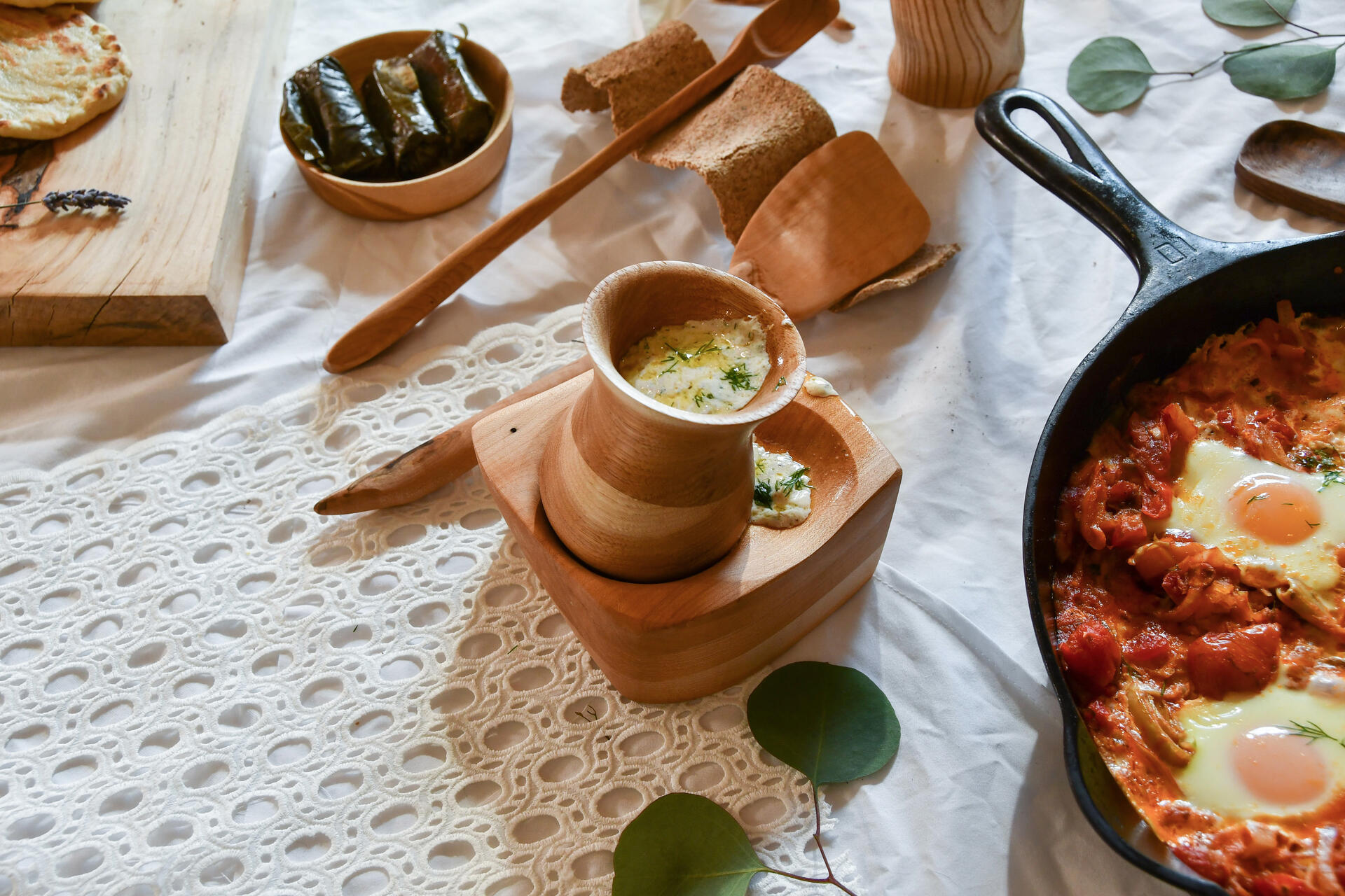 Wooden goblet filled with tzatziki and set into a larger wooden square bowl with two indents, wooden spoons, and part of a larger table spread on a white lace tablecloth. 