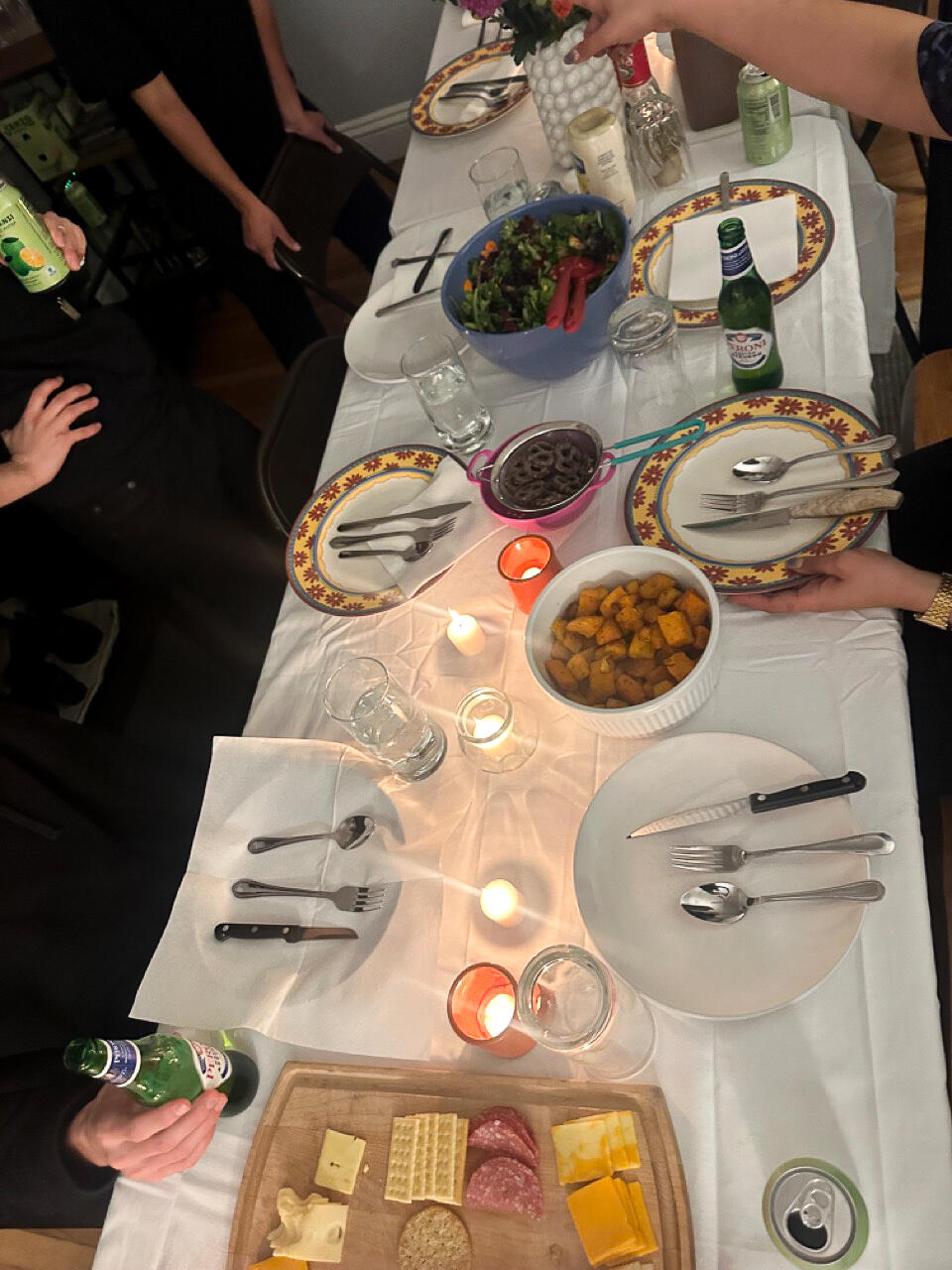 A long rectangular dinner table set with a white tablecloth, large round plates, and cutlery laid out in various patterns and orientations. 